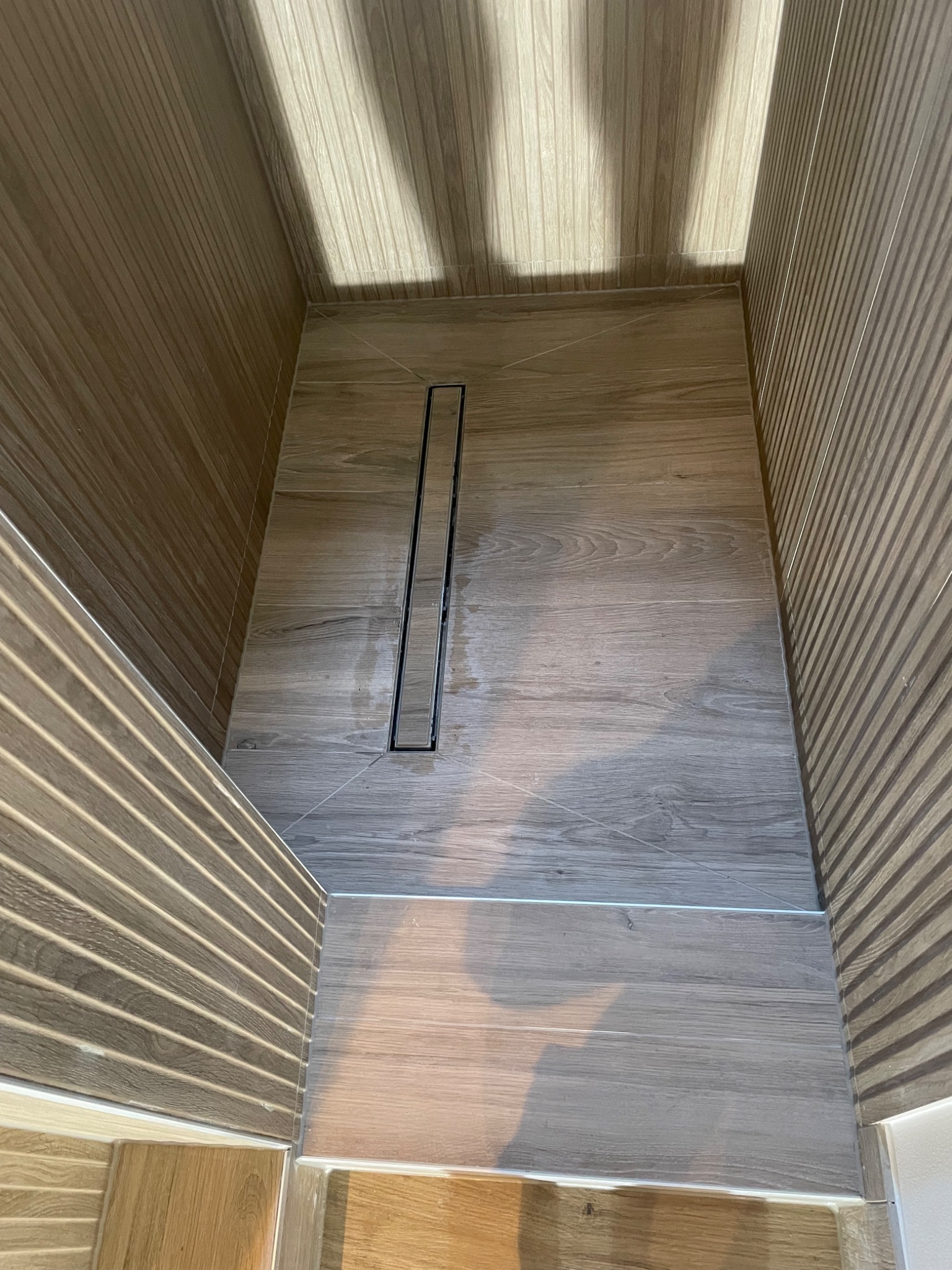 Wykończona kabina prysznicowa z odpływem liniowym, wyłożona płytkami imitującymi drewno na podłodze i ścianach z charakterystycznymi żłobieniami, oświetlona naturalnym światłem słonecznym...