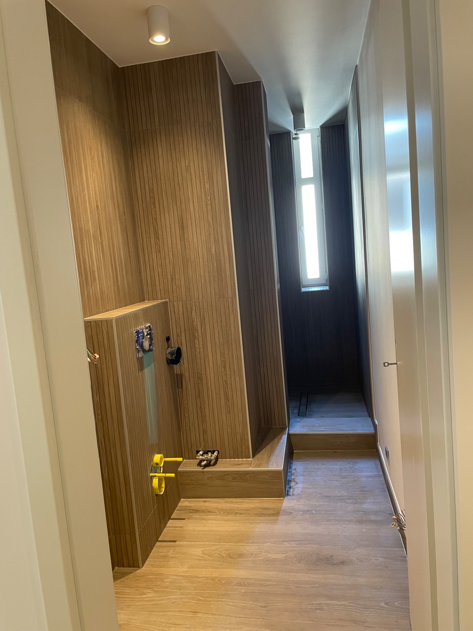 Wnętrze łazienki w trakcie remontu: drewnopodobne płytki na ścianach i podłodze, widoczne instalacje hydrauliczne, fragment okna w tle, surowy stan wykończenia.