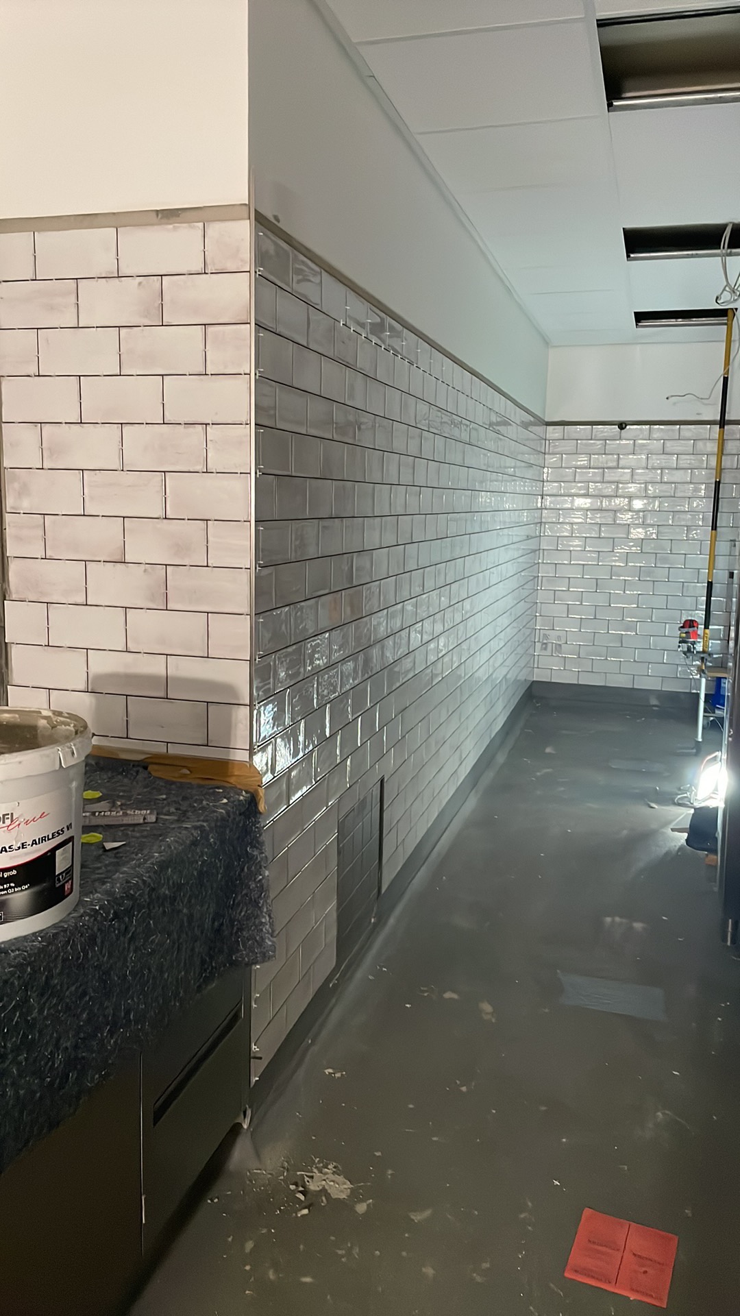 Układanie białych i szarych płytek typu metro na ścianach pomieszczenia, widoczny kubeł z klejem i resztki zaprawy na podłodze.