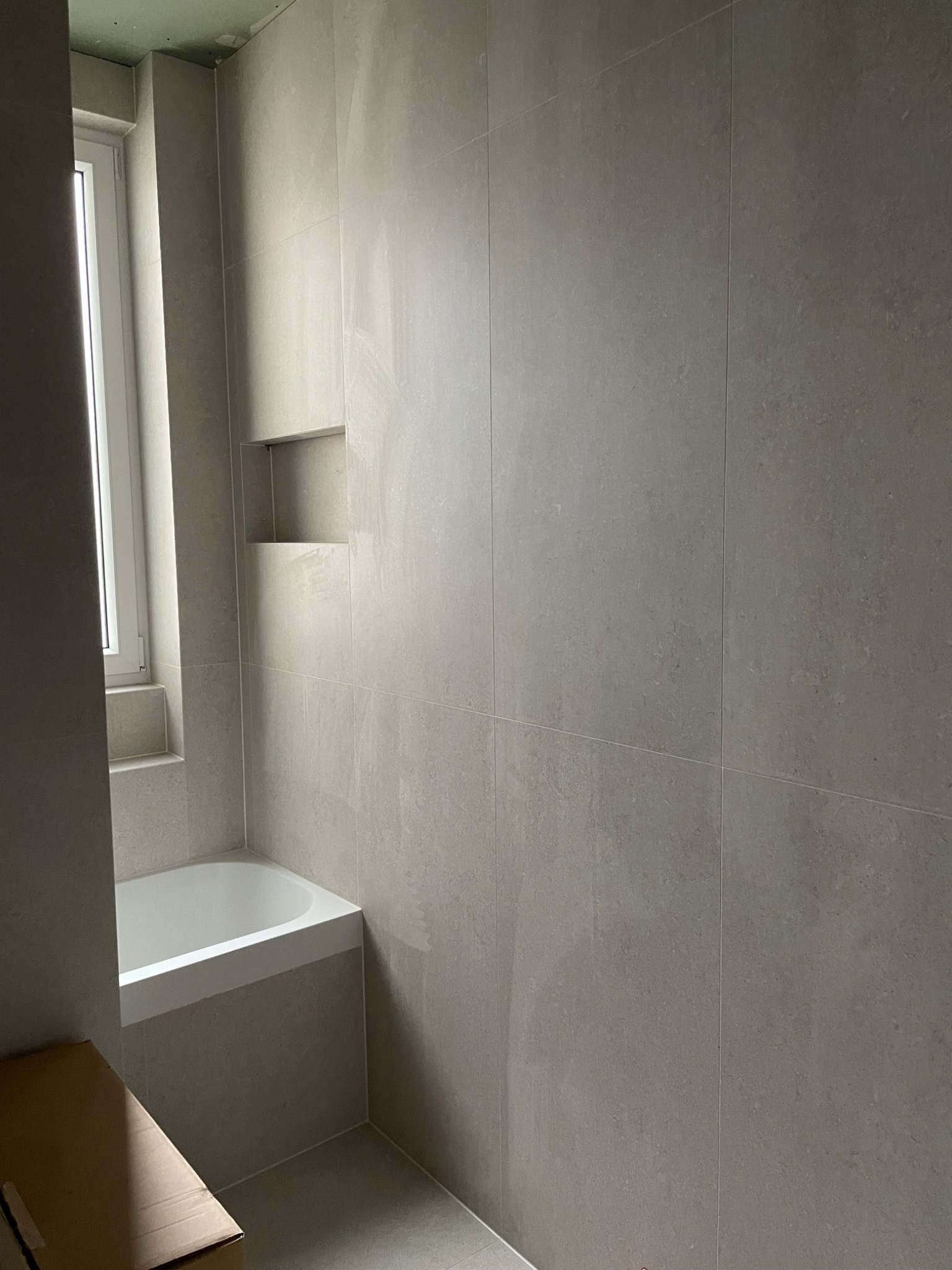 Nowoczesna łazienka w trakcie wykańczania, z wanną przy oknie i wnękami w szarej, matowej ścianie z płytek. Sufit jeszcze nie wykończony.