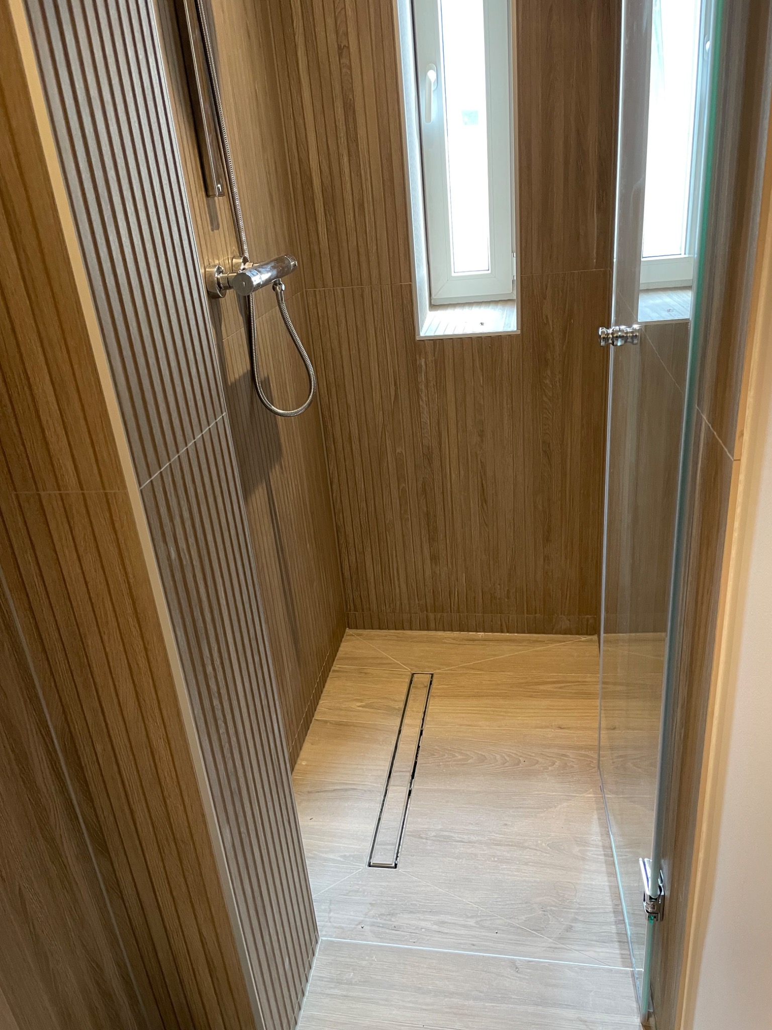 Nowoczesna kabina prysznicowa z odpływem liniowym, drewnopodobne płytki ścienne i podłogowe, okno w ramie PCV.