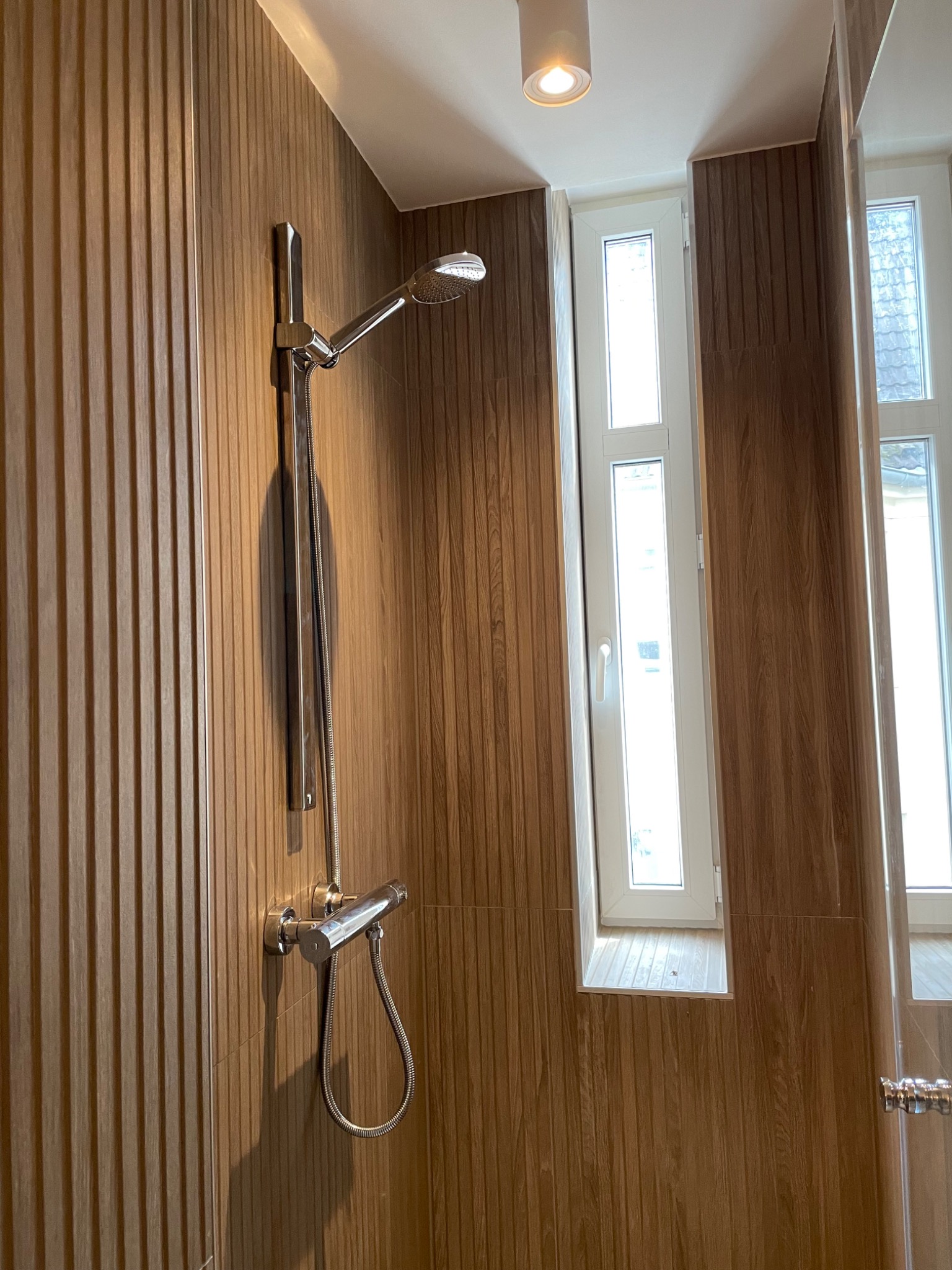 Nowoczesna kabina prysznicowa z panelem imitującym drewno, chromowana bateria z deszczownicą i wąskim oknem w białej ramie.