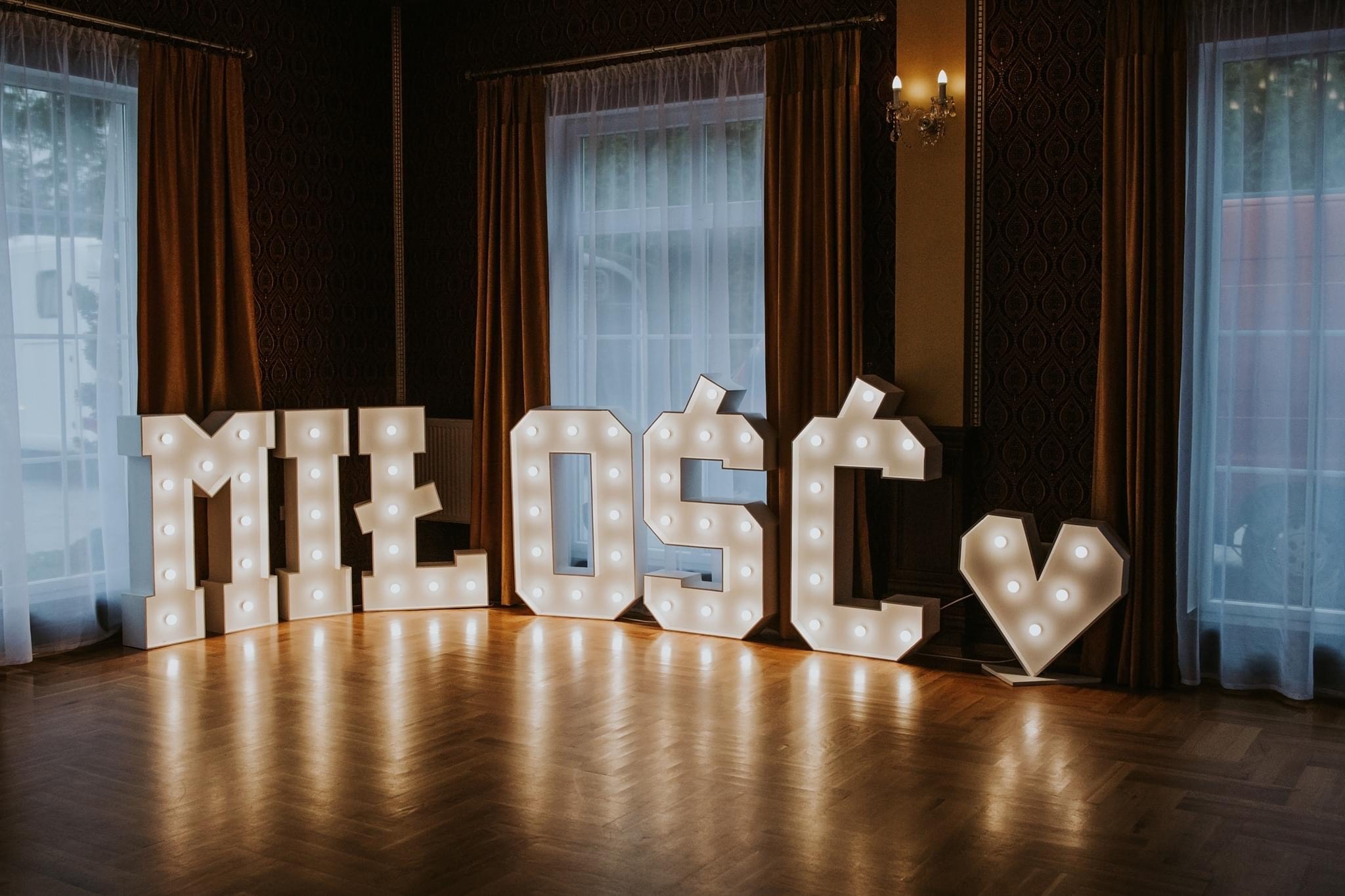 Podświetlany napis 'MIŁOŚĆ' z żarówkami w kształcie serca na drewnianej podłodze, ustawiony w eleganckim wnętrzu z zasłonami i tapetą w tle.