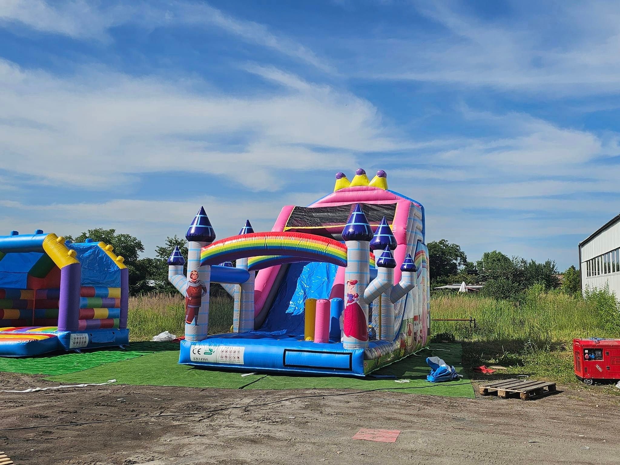 Dwa dmuchane zamki na trawie, jeden z motywem księżniczki i rycerza z tęczą, drugi kolorowy z tunelem, na tle błękitnego nieba z chmurami i budynku.