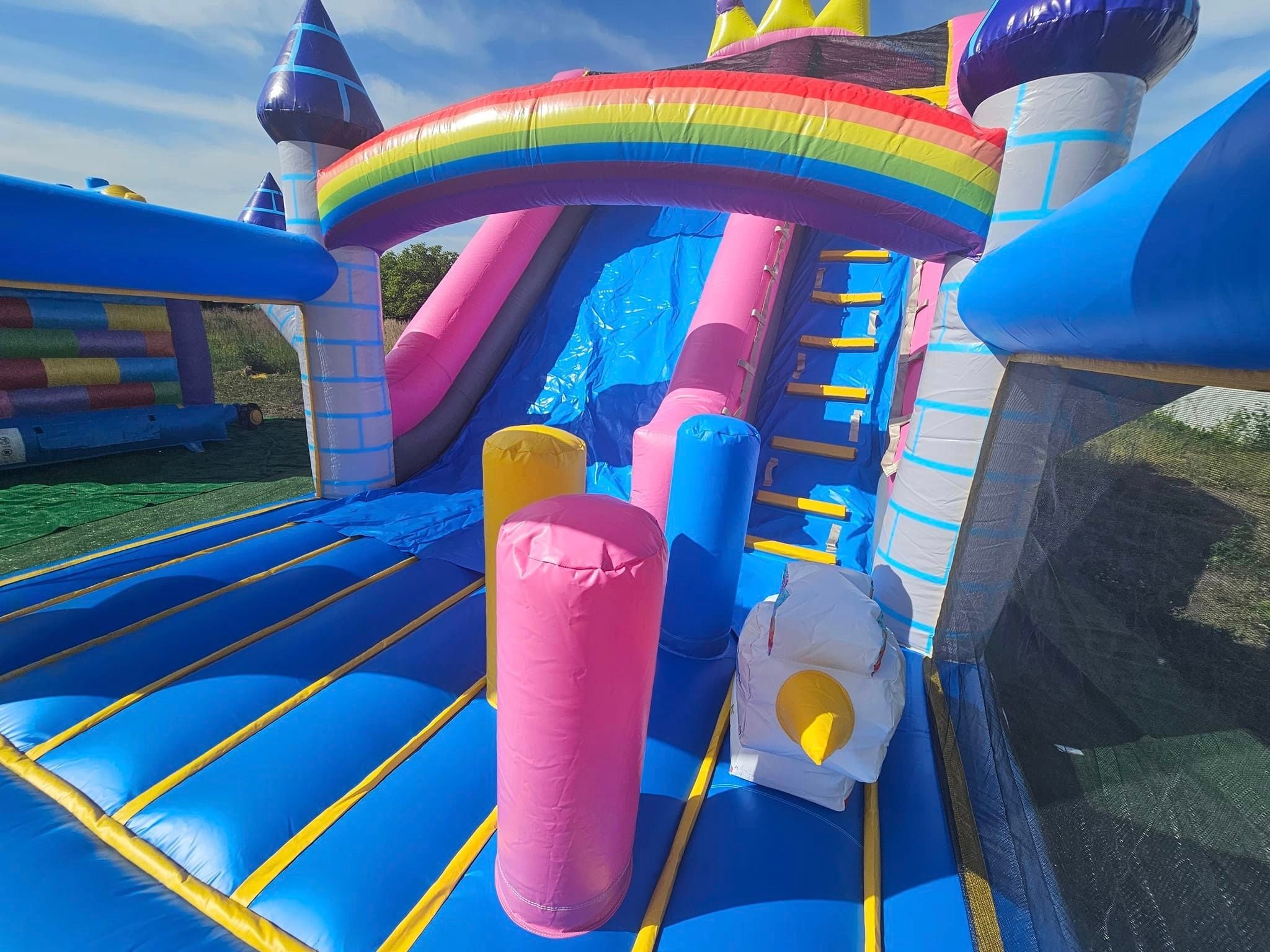 Kolorowy, dmuchany zamek ze zjeżdżalnią i tęczą, widok z dołu, w tle zielona trawa i błękitne niebo.