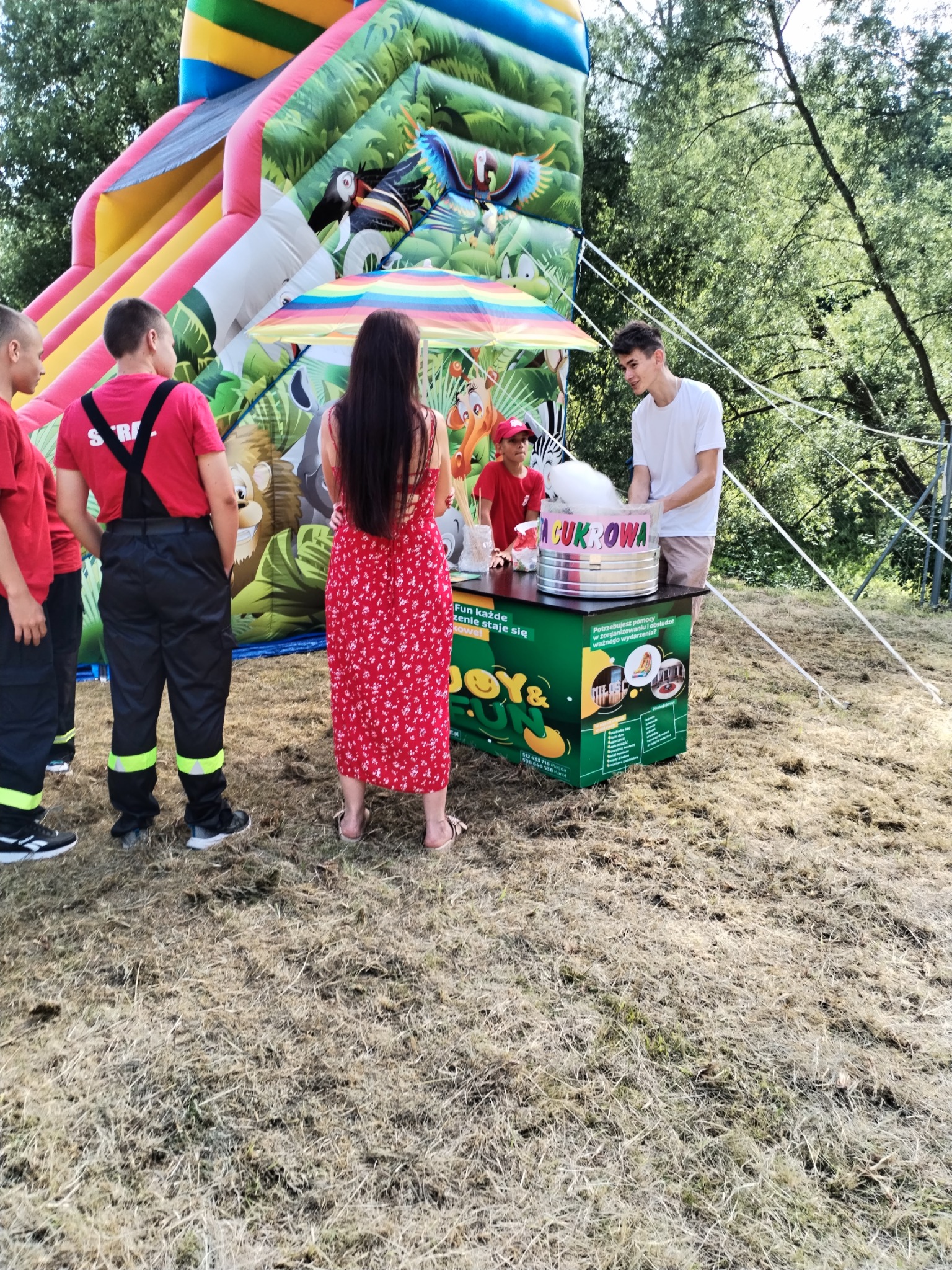 Obsługa waty cukrowej na tle dmuchanego zamku, widoczni ludzie czekający na swoją porcję, w tym grupa chłopców w strojach strażackich.