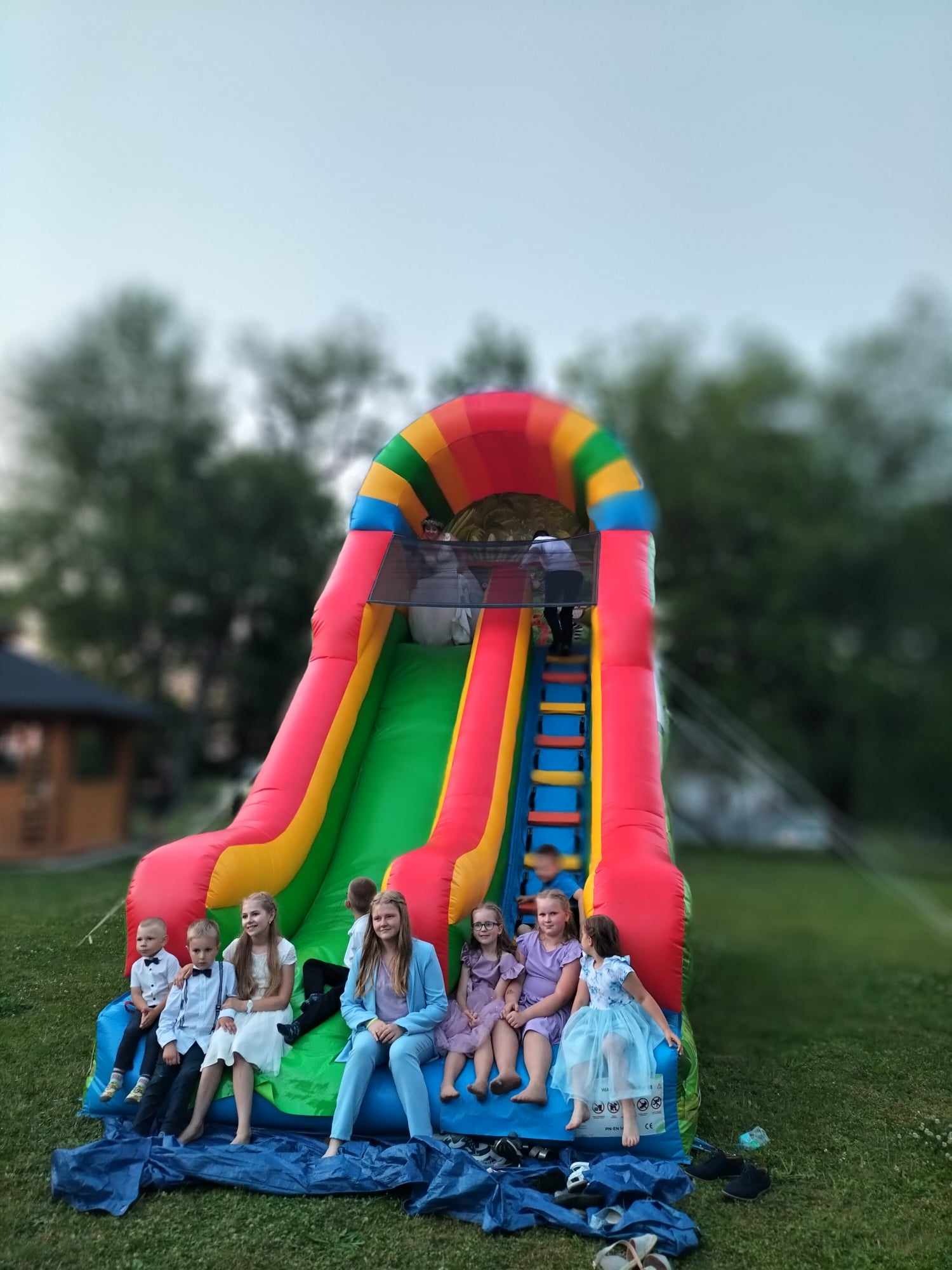 Grupa dzieci w eleganckich strojach siedzi przed dmuchaną zjeżdżalnią na zielonym trawniku, z osobą wchodzącą na górę po schodach.