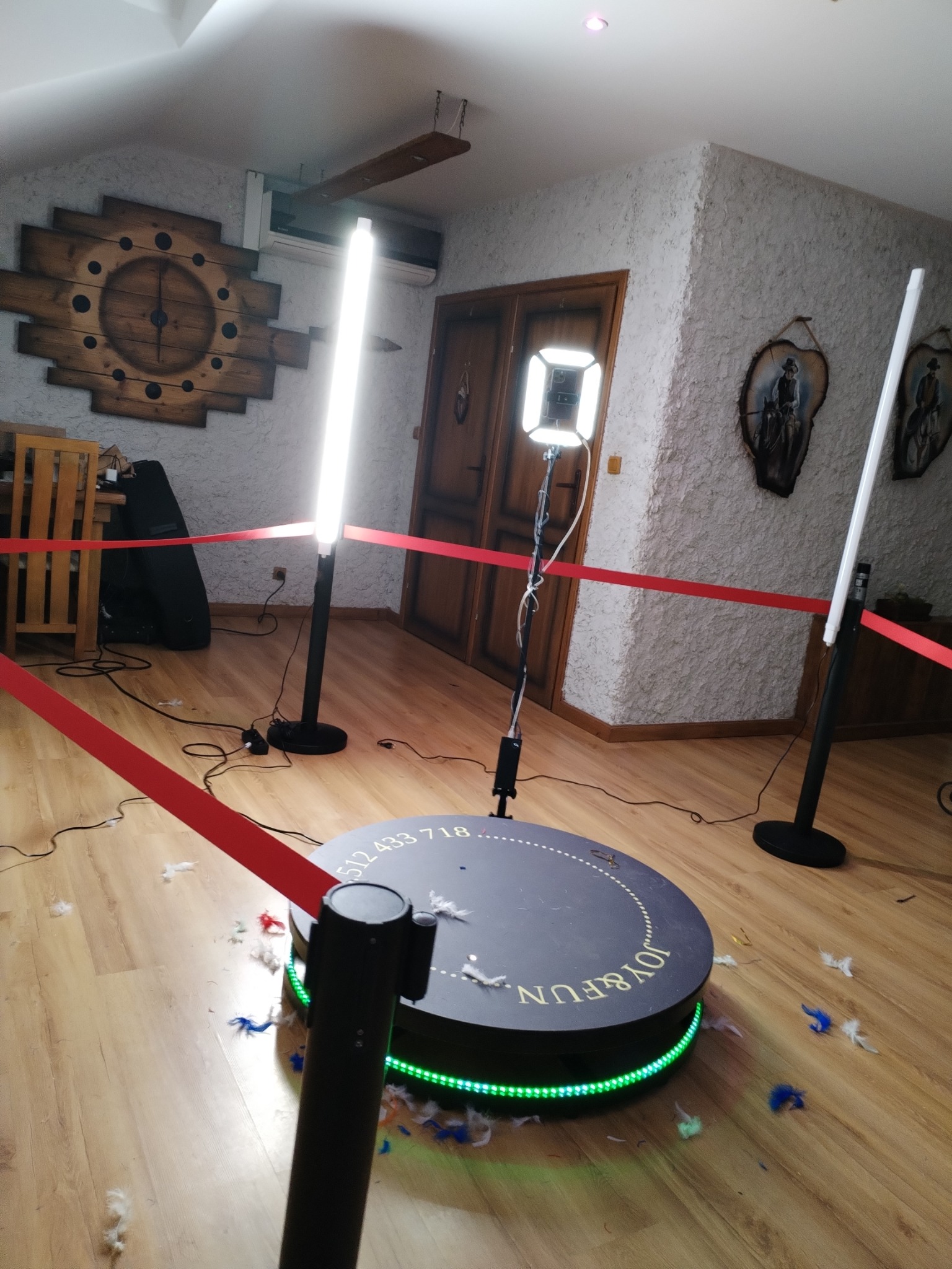 Okrągła platforma obrotowa z napisem 'JOY & FUN' otoczona zielonymi diodami LED i rozrzuconymi piórami, ograniczona czerwonymi taśmami, oświetlona lampami LED, w tle rustykalne wnętrze z drewnianym...