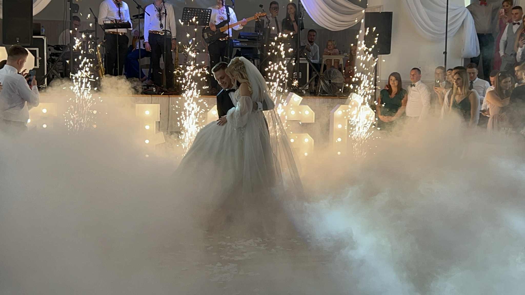 Pierwszy taniec pary młodej w dymie i blasku fontann iskier na tle zespołu muzycznego i gości weselnych.