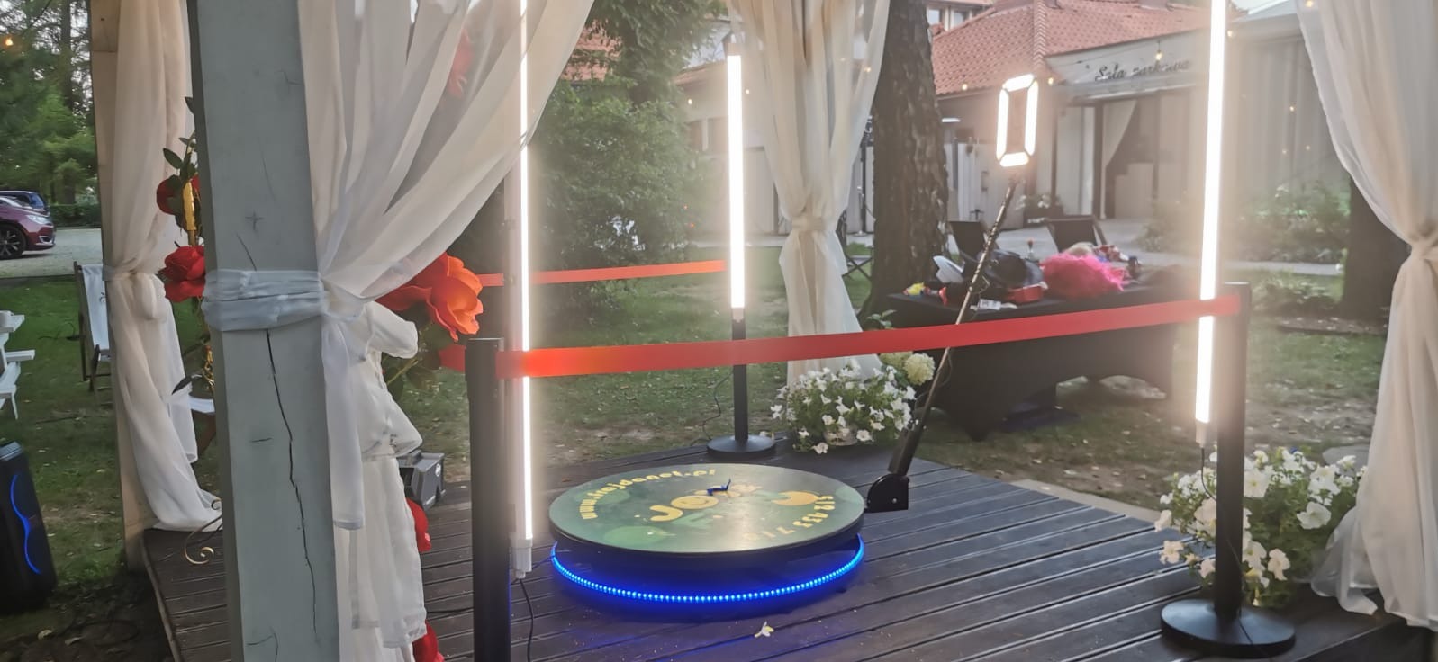 Ozdobiona kwiatami i białymi tkaninami fotobudka 360 na podeście z desek, z oświetleniem LED, otoczona zielenią. W tle budynek z napisem 'Sala bankietowa'.