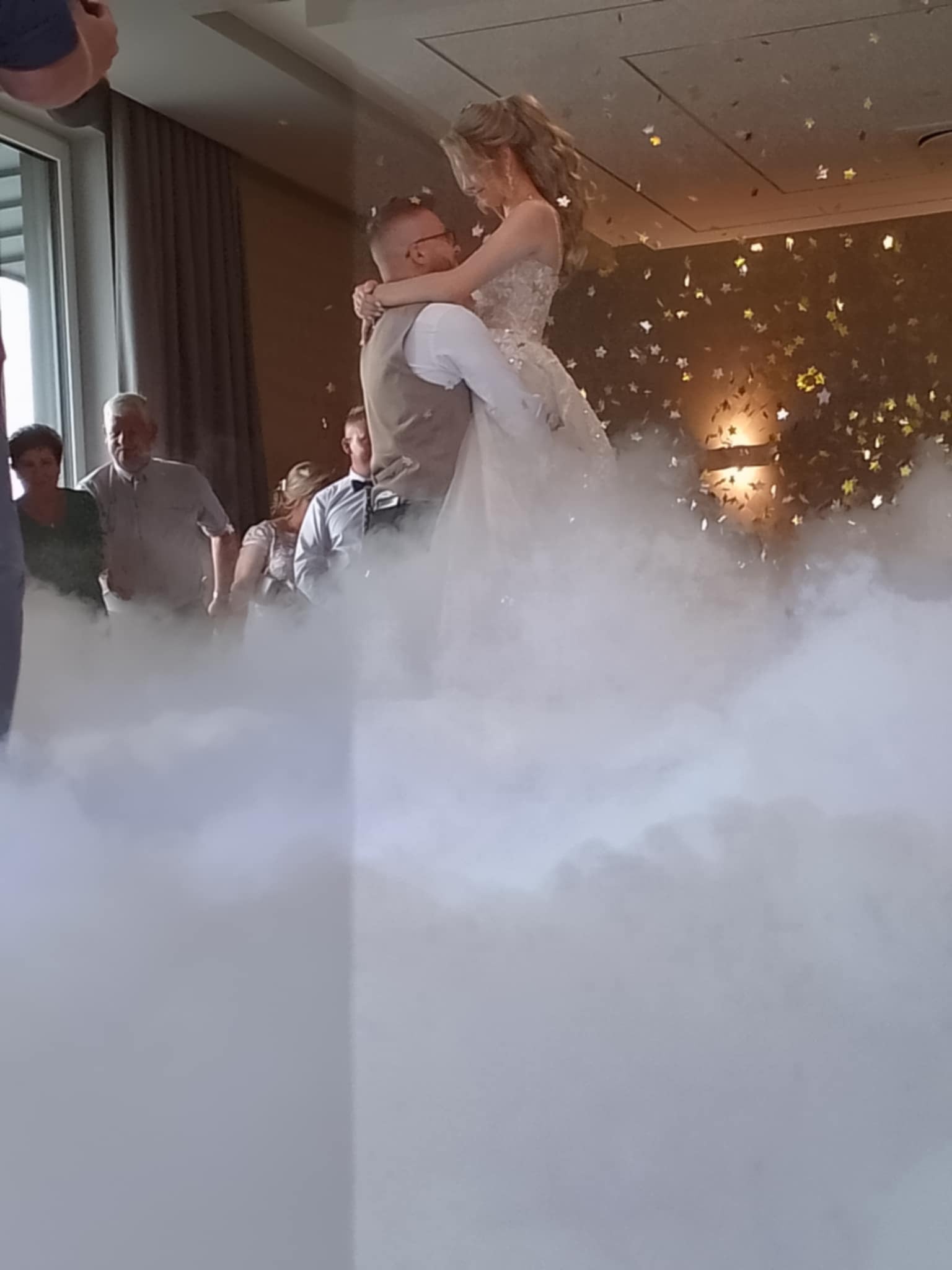 Para młoda tańczy w chmurze dymu i strugach konfetti podczas wesela. W tle goście weselni i elementy dekoracji sali.