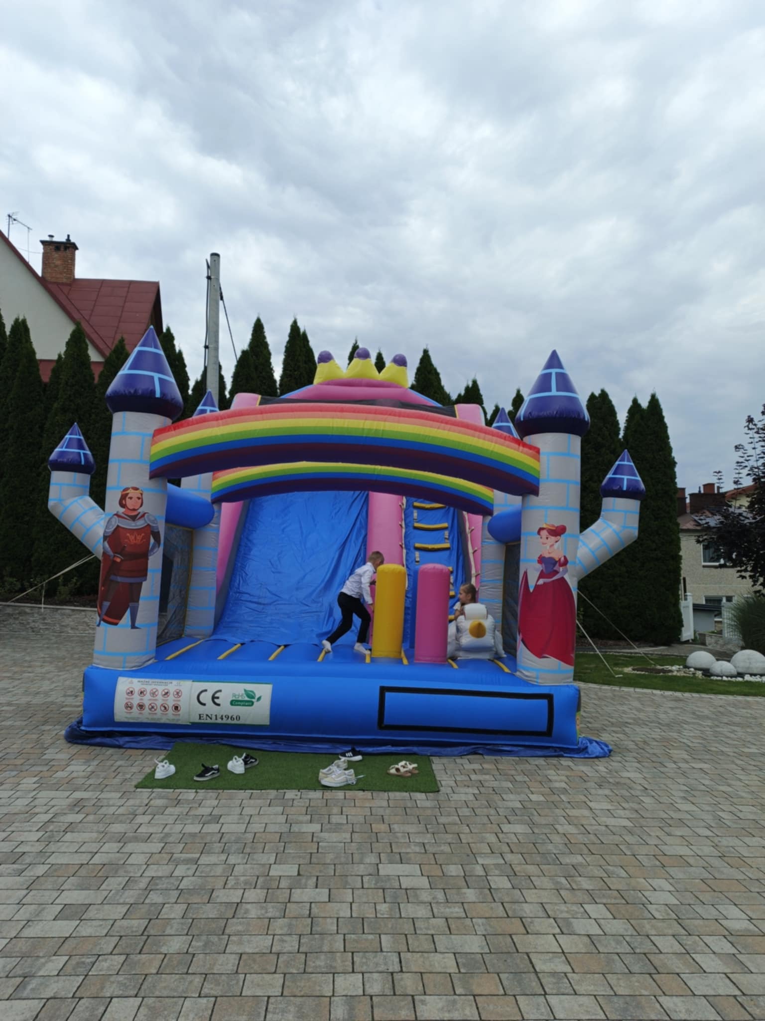 Kolorowy, dmuchany zamek z motywem rycerza i księżniczki, zjeżdżalnią i tęczą. Dzieci korzystają z atrakcji na tle brukowanego podwórza i zielonych drzew.