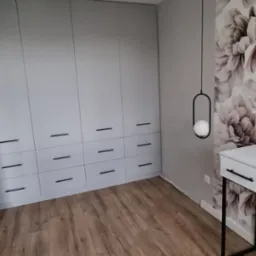 Jasnoszara szafa na wymiar z czarnymi uchwytami, drewniana podłoga, toaletka z czarnymi nogami i kwiatowa tapeta w tle, nowoczesna lampa wisząca.