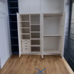 Biała szafa wnękowa z otwartymi drzwiami, widoczne półki i szuflady, z boku regał w kolorze granatowym, na podłodze niebieska taśma w kształcie X.