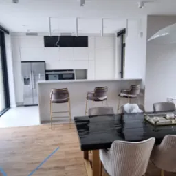 Nowoczesna kuchnia z białymi szafkami, wyspą z hokerami i czarnym stołem z krzesłami w trakcie wykańczania, widoczne niebieskie taśmy na podłodze.