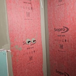 RENOVHOUM - Przygotowanie do hydroizolacji w narożniku łazienki: ściany pokryte czerwoną membraną Sopro AEB 640, widoczne wyprowadzenia instalacji wodnej, sufit z płyt kartonowo-gipsowych, podłoga z OSB...