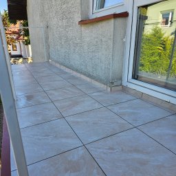 RENOVHOUM - Wyłożony beżowymi płytkami taras przy budynku z oknem, widok na zewnątrz z odbiciem zieleni w szybie.