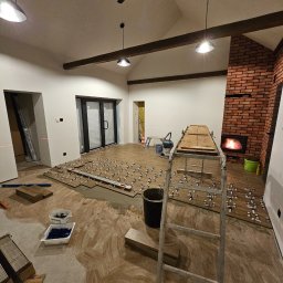 RENOVHOUM - Układanie płytek podłogowych imitujących drewno w przestronnym salonie z ceglanym kominkiem, widoczne poziomice, krzyżyki dystansowe i drabina.