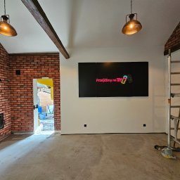 RENOVHOUM - Wnętrze w trakcie prac wykończeniowych: surowy beton na podłodze, ceglana ściana z kominkiem, biała ściana z neonowym napisem i rusztowanie.