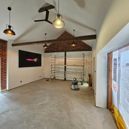 RENOVHOUM - Wnętrze w trakcie remontu: surowy beton na podłodze, ściany częściowo pomalowane na biało, fragment ściany z cegły, drewniane belki stropowe, wiszące lampy w stylu industrialnym, drabina...