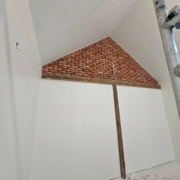 RENOVHOUM - Skośna ściana z fragmentem dekoracyjnej cegły na poddaszu, widoczne drewniane belki, białe ściany, kabel elektryczny zwisający z sufitu i fragment rusztowania.