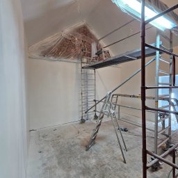 RENOVHOUM - Wnętrze w trakcie malowania, ściany świeżo pomalowane na biało, rusztowanie w pomieszczeniu, zabezpieczona folią ceglana ściana pod skosem sufitu, widoczne lampy budowlane.