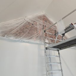 RENOVHOUM - Przygotowanie do malowania ściany z cegieł w Kunowie, zabezpieczonej folią, widoczna drabina rusztowania i świeżo pomalowana biała ściana.