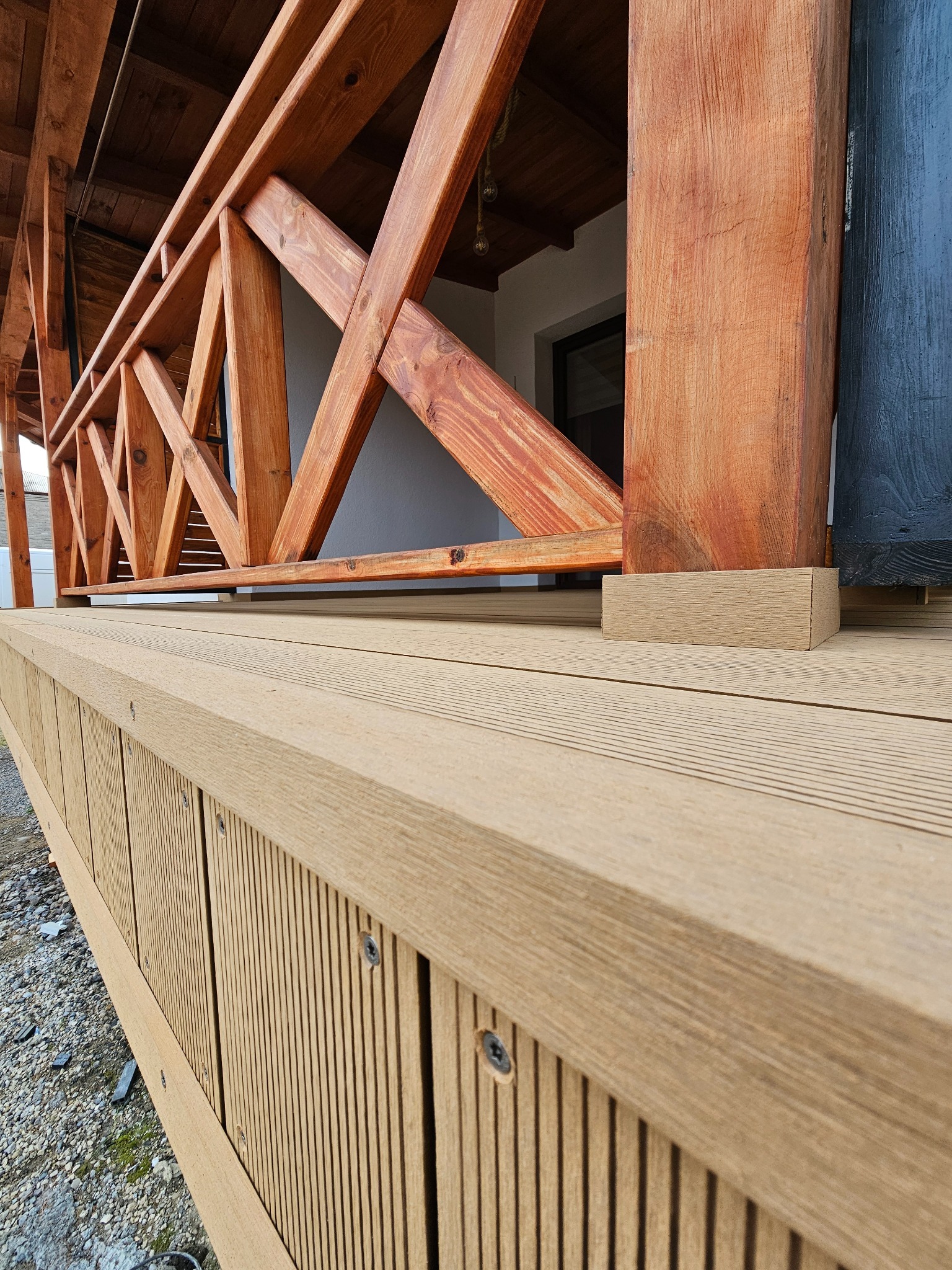 Drewniana konstrukcja tarasu z widocznymi poziomymi deskami kompozytowymi, balustradą z krzyżujących się belek oraz masywnymi słupami nośnymi w ciepłym odcieniu brązu.