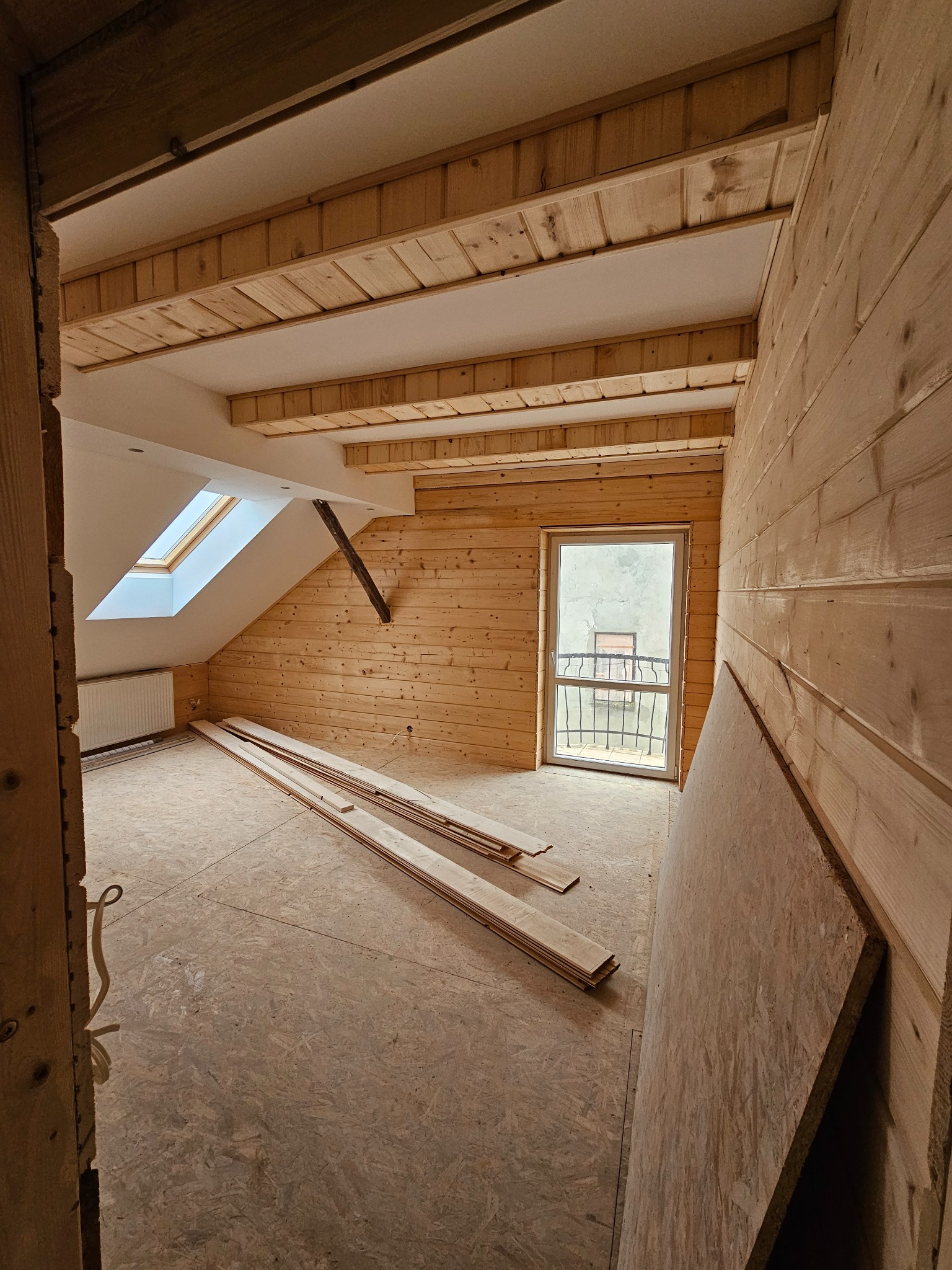 Poddasze w trakcie wykańczania drewnem: boazeria na ścianach i suficie, okno dachowe, deski na podłodze, płyta OSB oparta o ścianę.