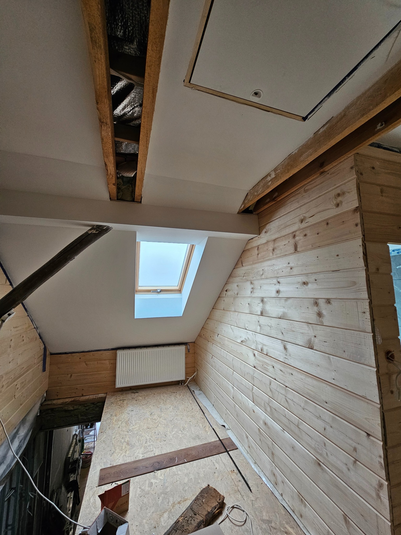 Poddasze w trakcie wykańczania, widoczne drewniane panele ścienne, okno dachowe, grzejnik, fragment sufitu z otworem rewizyjnym i odsłoniętą konstrukcją dachu z izolacją.