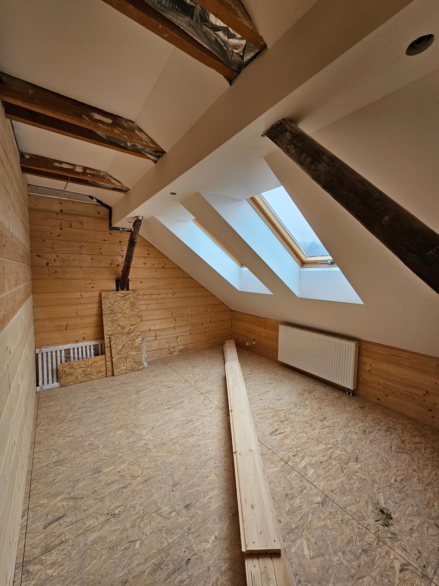 Poddasze w trakcie prac wykończeniowych: drewniane panele ścienne i podłogowe OSB, trzy okna dachowe, grzejnik, widoczne elementy konstrukcyjne dachu.