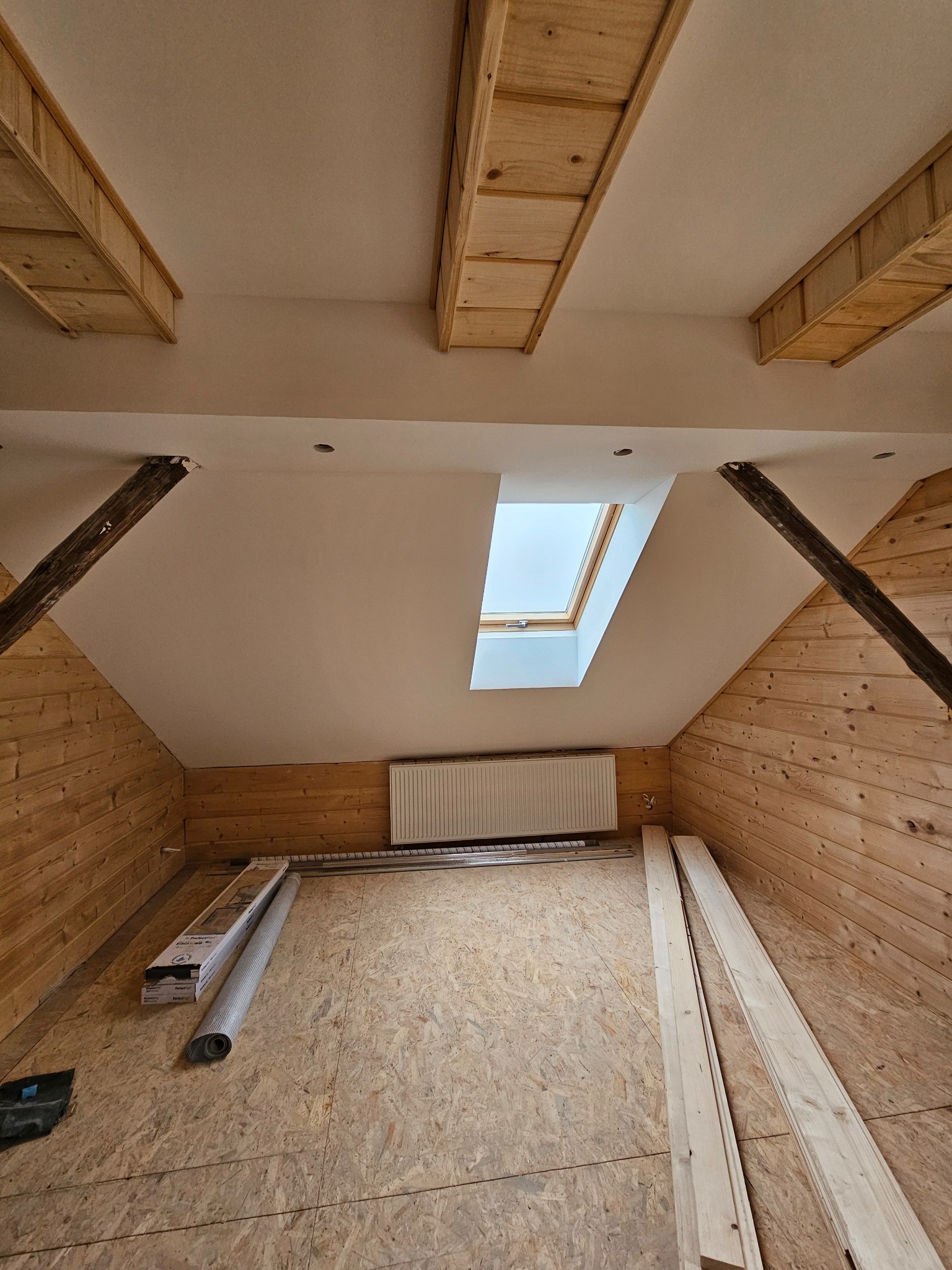 Poddasze w trakcie prac wykończeniowych: drewniana boazeria, okno dachowe, widoczne drewniane belki stropowe, grzejnik oraz materiały budowlane na podłodze z płyty OSB.