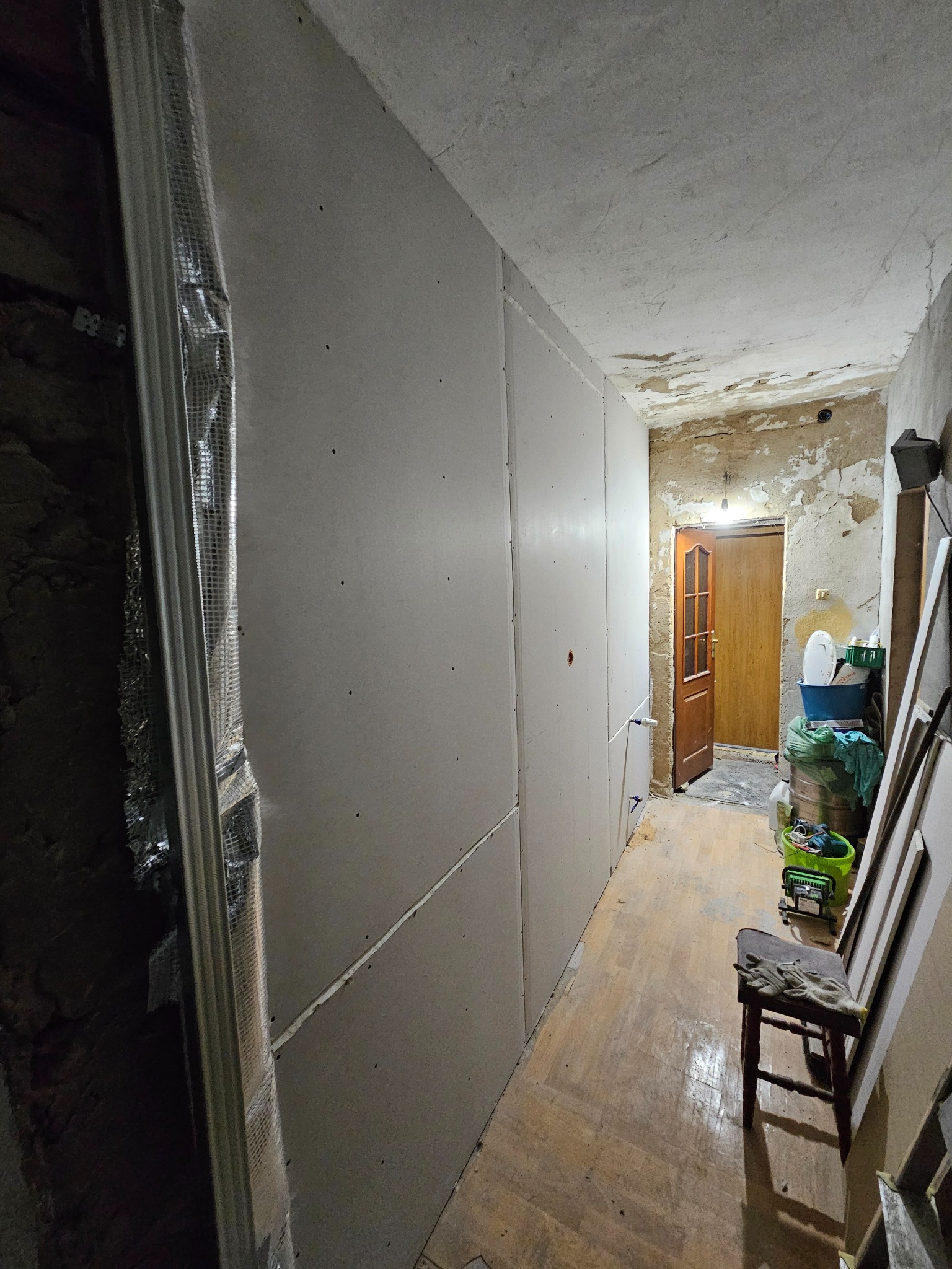 Korytarz w trakcie prac: ściany wyłożone płytami gipsowo-kartonowymi, widoczne profile montażowe, surowe ściany w tle, drewniana podłoga, krzesło z rękawicami roboczymi.