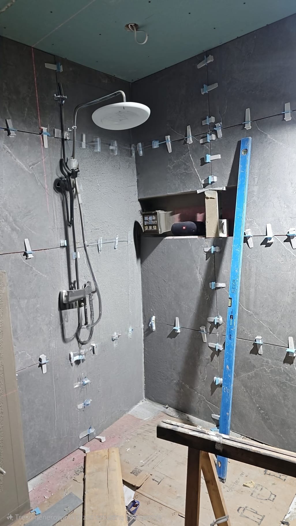 Wnętrze łazienki w trakcie układania grafitowych płytek imitujących kamień, widoczny prysznic z deszczownicą, poziomica oparta o ścianę i system poziomowania płytek z krzyżykami dystansowymi, wnęka...