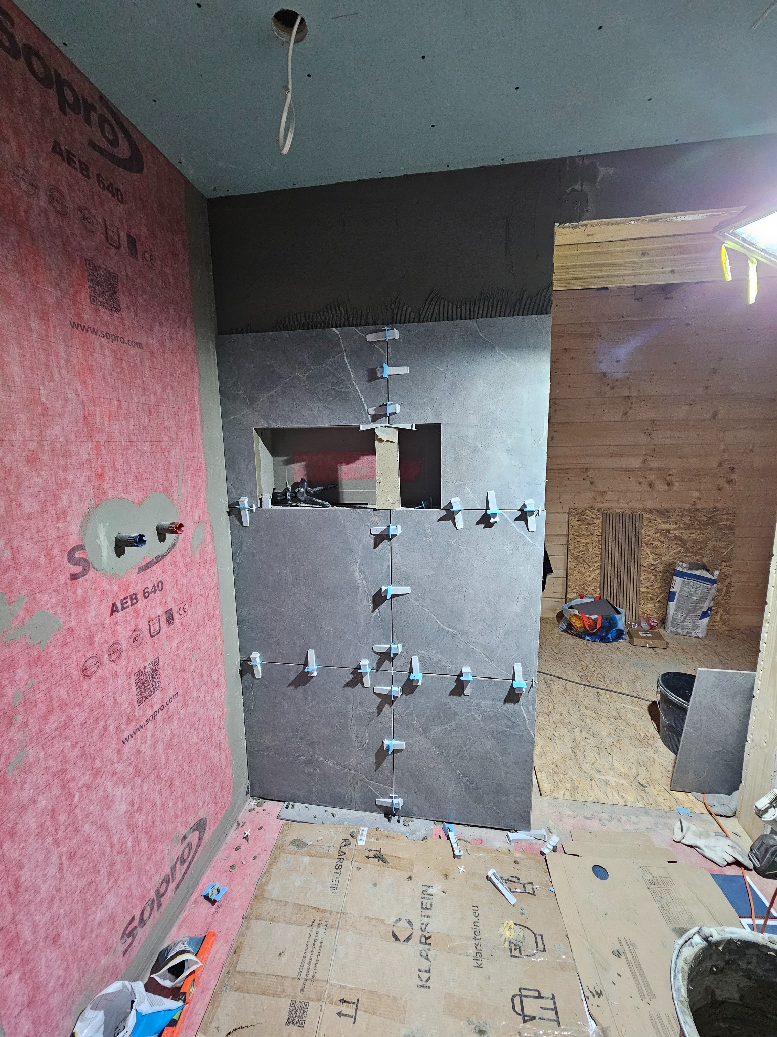 Montaż ciemnoszarych płytek na ścianie w łazience, widoczne krzyżyki dystansowe, hydroizolacja Sopro na sąsiedniej ścianie, przygotowane otwory na armaturę i narzędzia na podłodze wyłożonej kartonem.