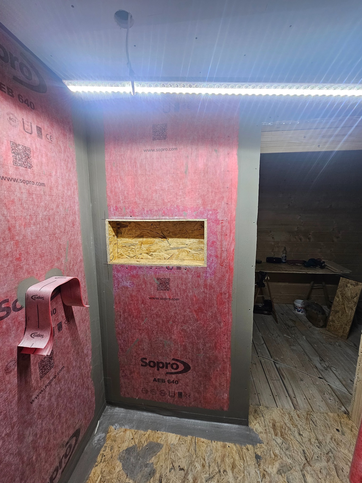 Przygotowanie do remontu łazienki: ściany wyłożone wodoodporną membraną Sopro AEB 640, wnęka w ścianie z surowego drewna, oświetlenie LED na suficie, fragment podłogi z płyt OSB.