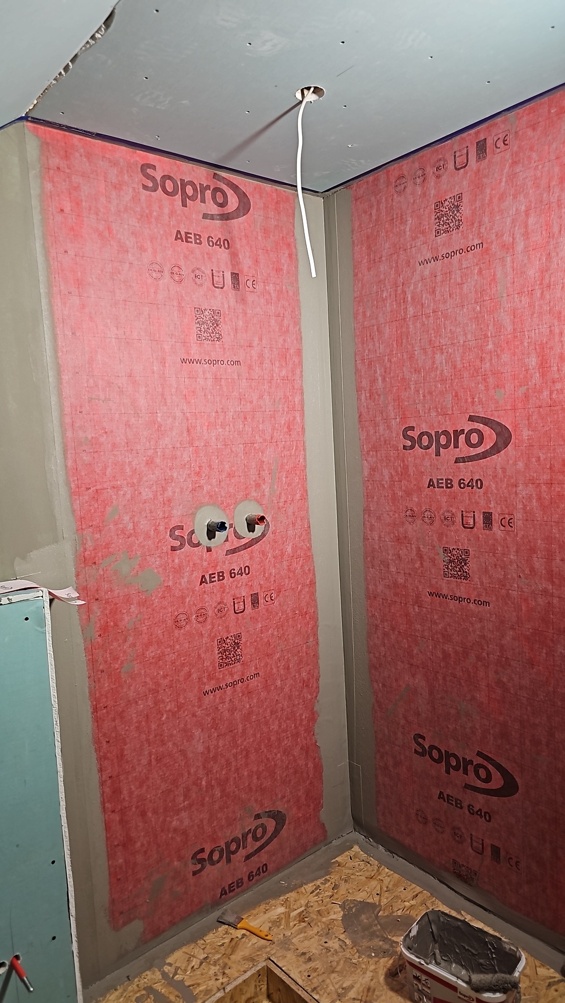 Przygotowanie do hydroizolacji w narożniku łazienki: ściany pokryte czerwoną membraną Sopro AEB 640, widoczne wyprowadzenia instalacji wodnej, sufit z płyt kartonowo-gipsowych, podłoga z OSB...