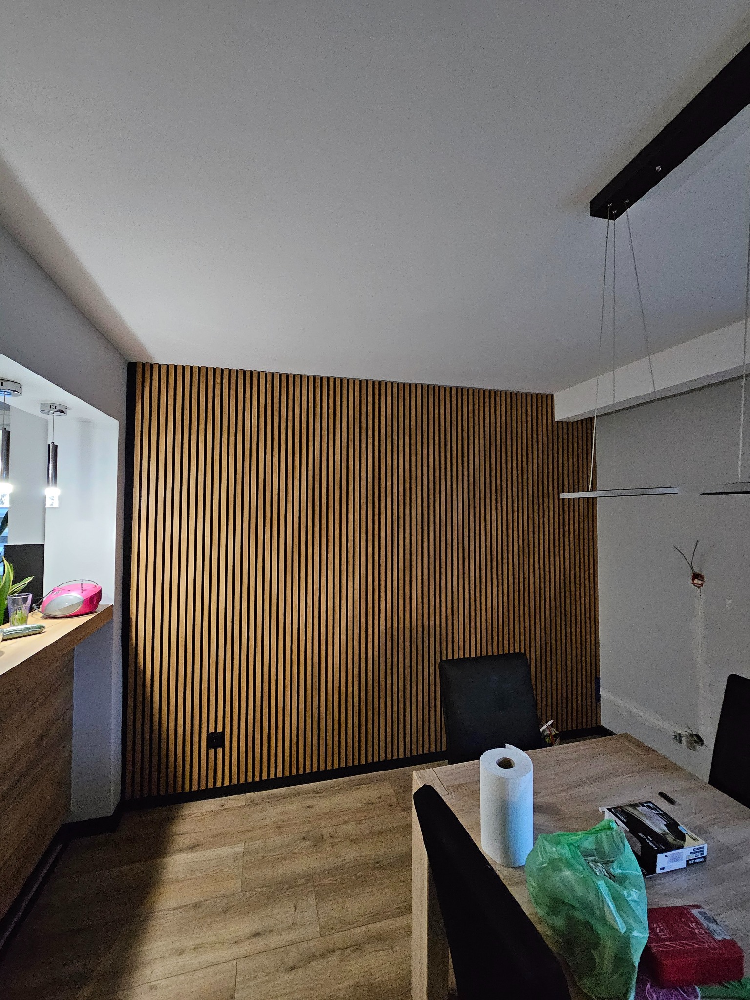 Panel ścienny z drewnianych listewek w salonie, z widocznym stołem, krzesłami i lampą wiszącą; na stole rolka papieru i torba foliowa.