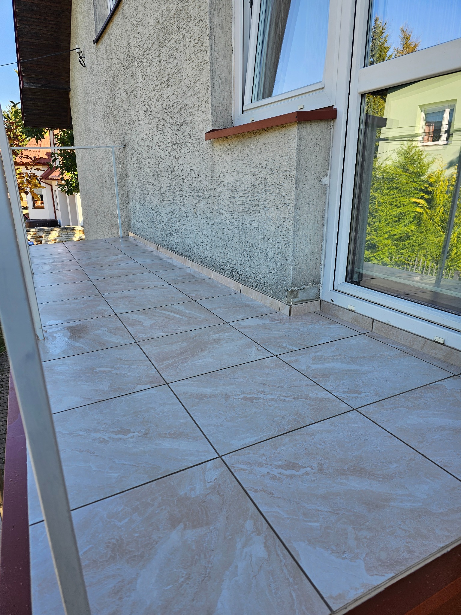 Wyłożony beżowymi płytkami taras przy budynku z oknem, widok na zewnątrz z odbiciem zieleni w szybie.