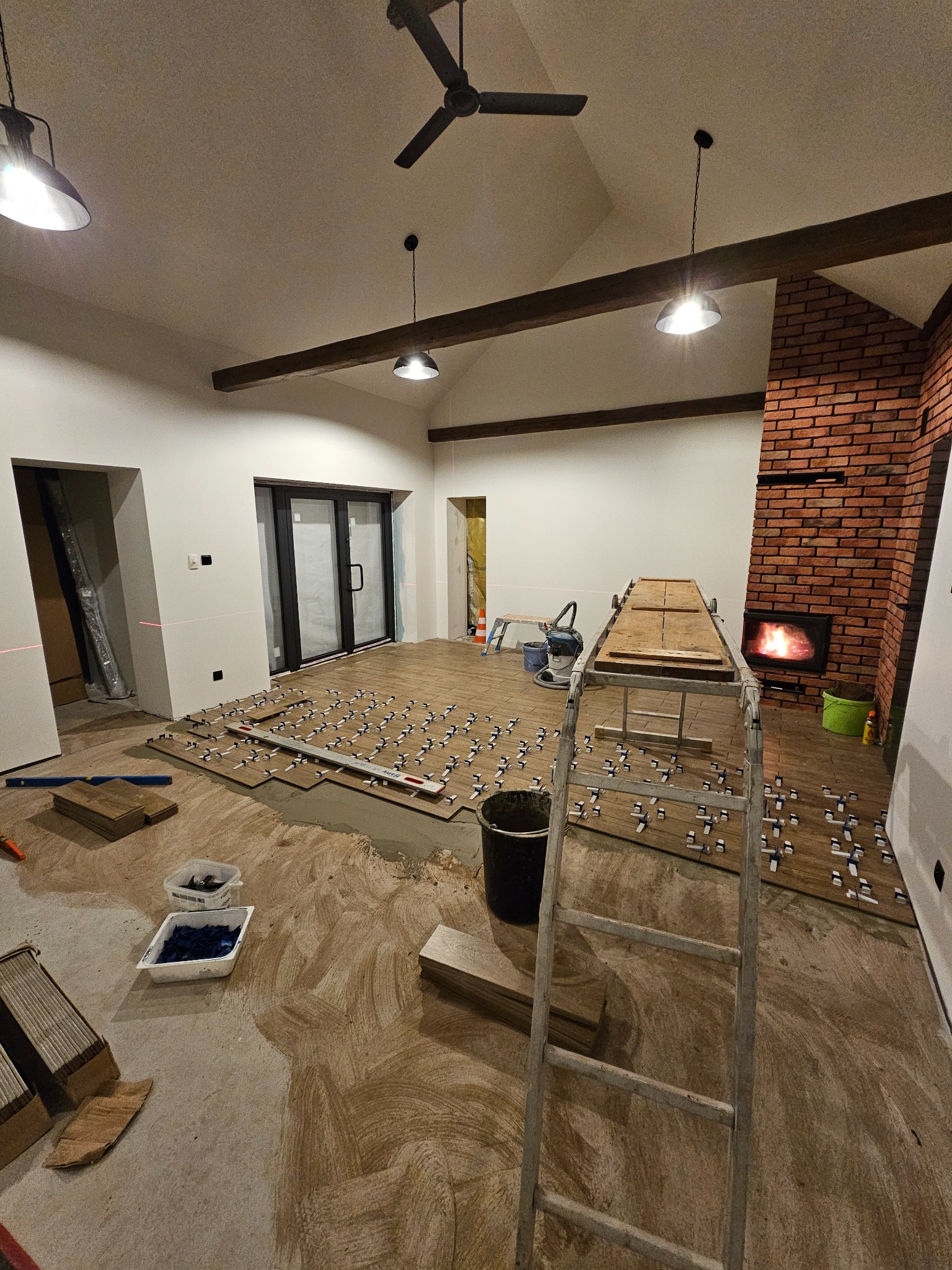 Układanie płytek podłogowych imitujących drewno w przestronnym salonie z ceglanym kominkiem, widoczne poziomice, krzyżyki dystansowe i drabina.