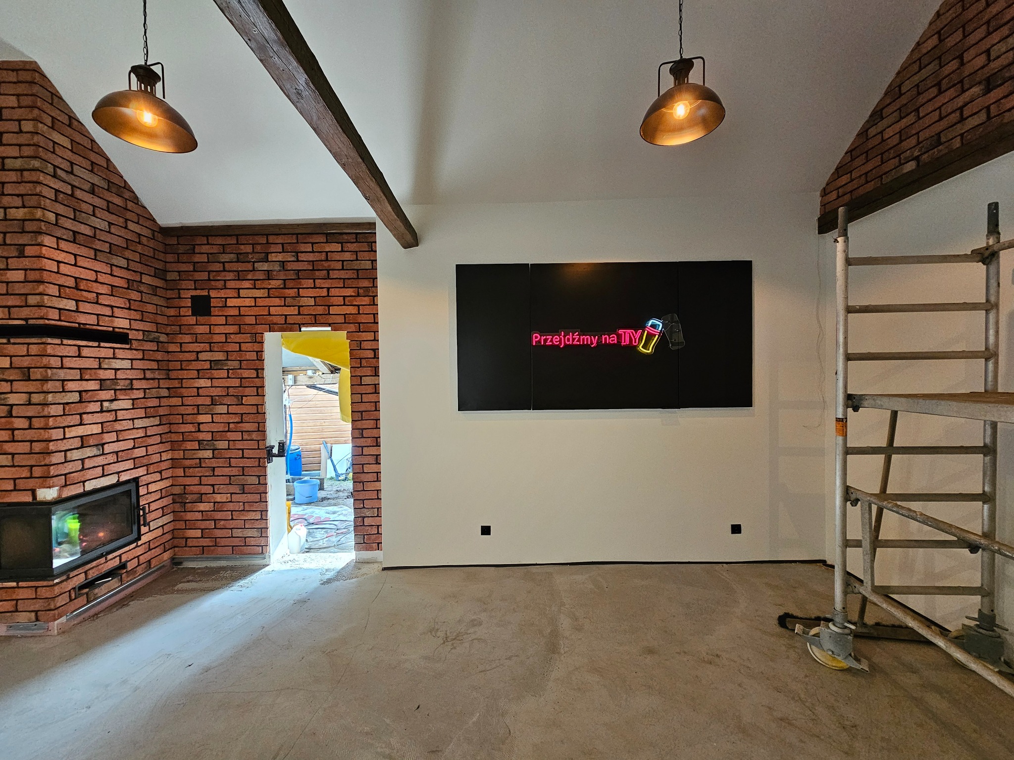 Wnętrze w trakcie prac wykończeniowych: surowy beton na podłodze, ceglana ściana z kominkiem, biała ściana z neonowym napisem i rusztowanie.