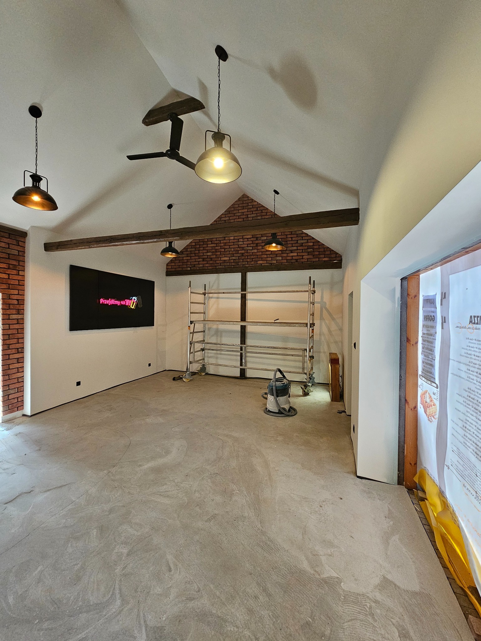 Wnętrze w trakcie remontu: surowy beton na podłodze, ściany częściowo pomalowane na biało, fragment ściany z cegły, drewniane belki stropowe, wiszące lampy w stylu industrialnym, drabina...