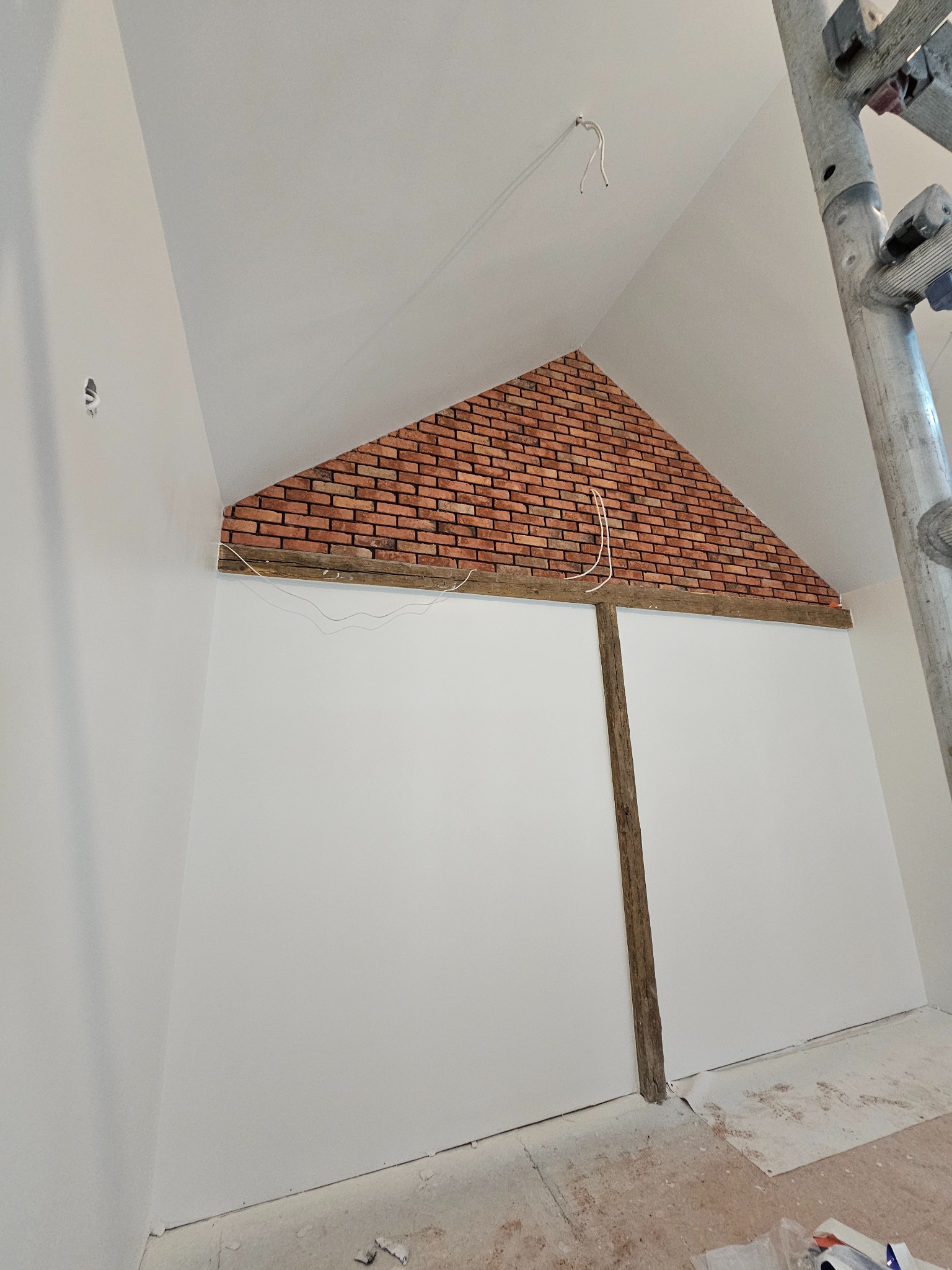 Skośna ściana z fragmentem dekoracyjnej cegły na poddaszu, widoczne drewniane belki, białe ściany, kabel elektryczny zwisający z sufitu i fragment rusztowania.
