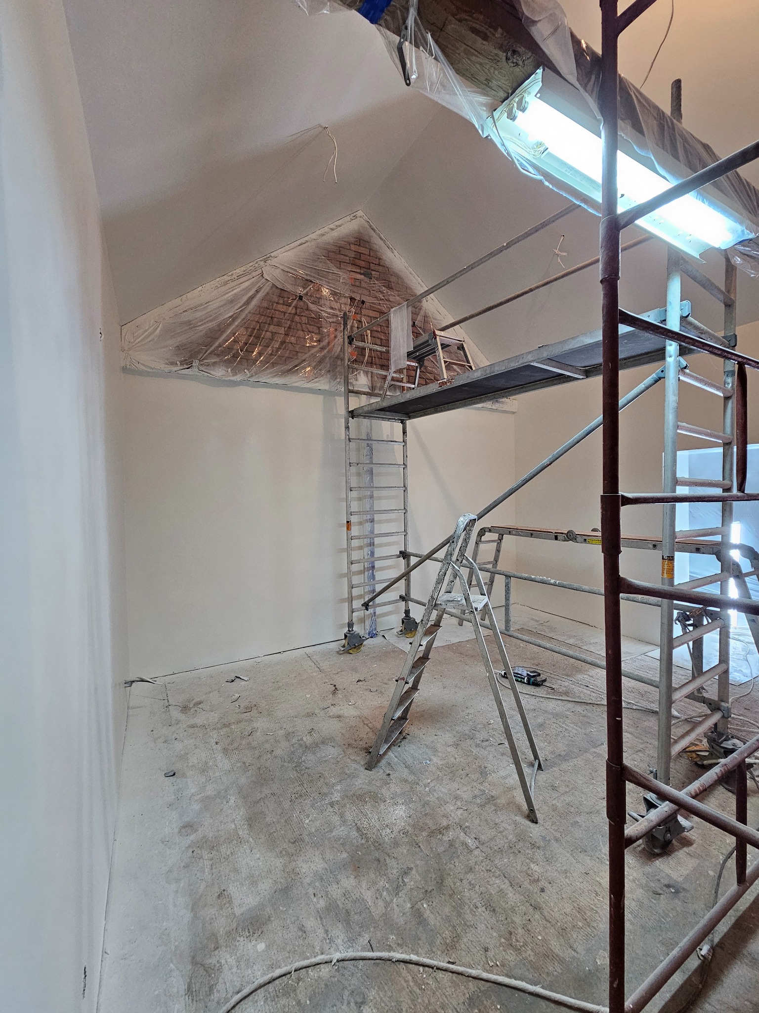 Wnętrze w trakcie malowania, ściany świeżo pomalowane na biało, rusztowanie w pomieszczeniu, zabezpieczona folią ceglana ściana pod skosem sufitu, widoczne lampy budowlane.