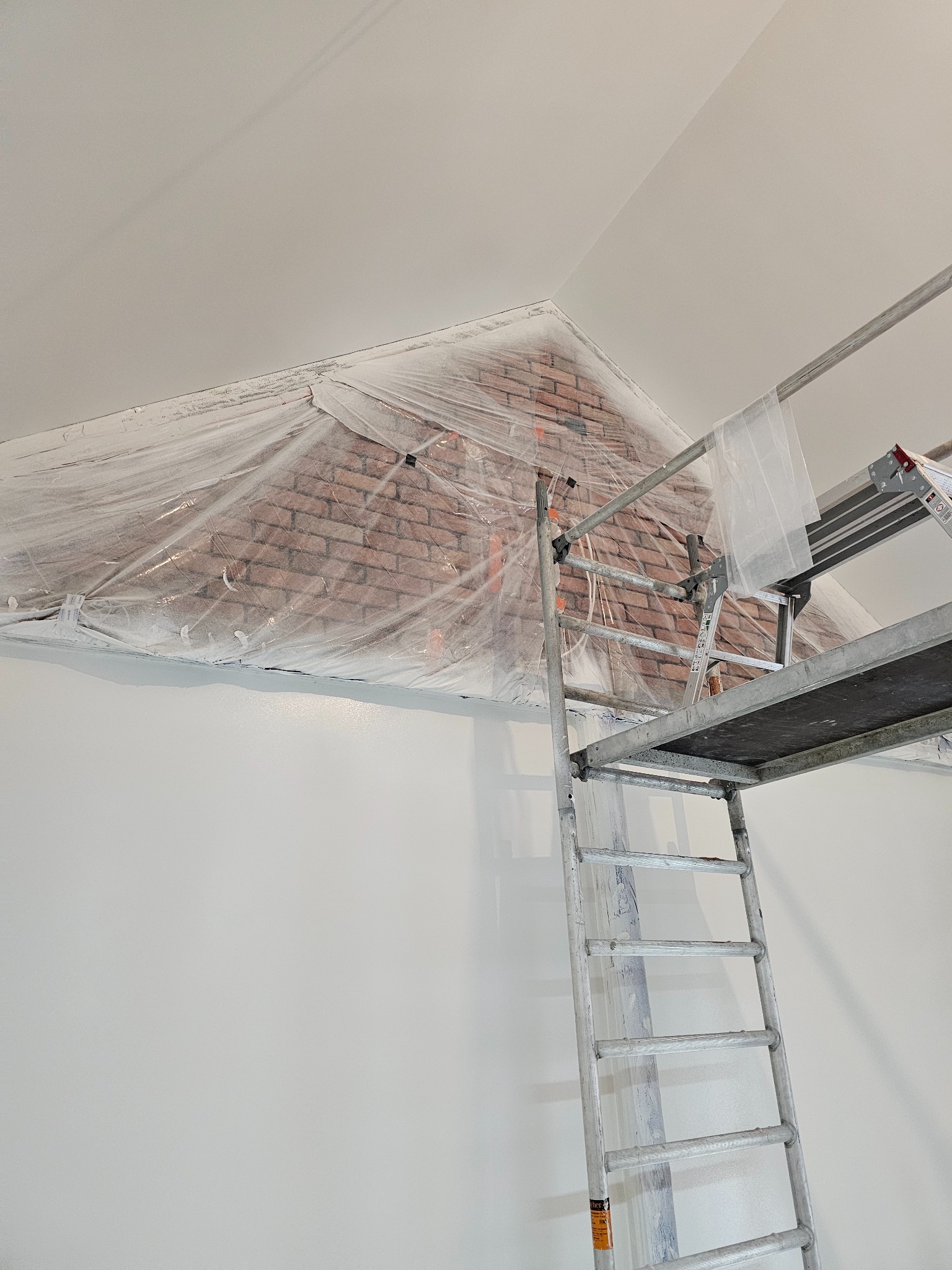 Przygotowanie do malowania ściany z cegieł w Kunowie, zabezpieczonej folią, widoczna drabina rusztowania i świeżo pomalowana biała ściana.