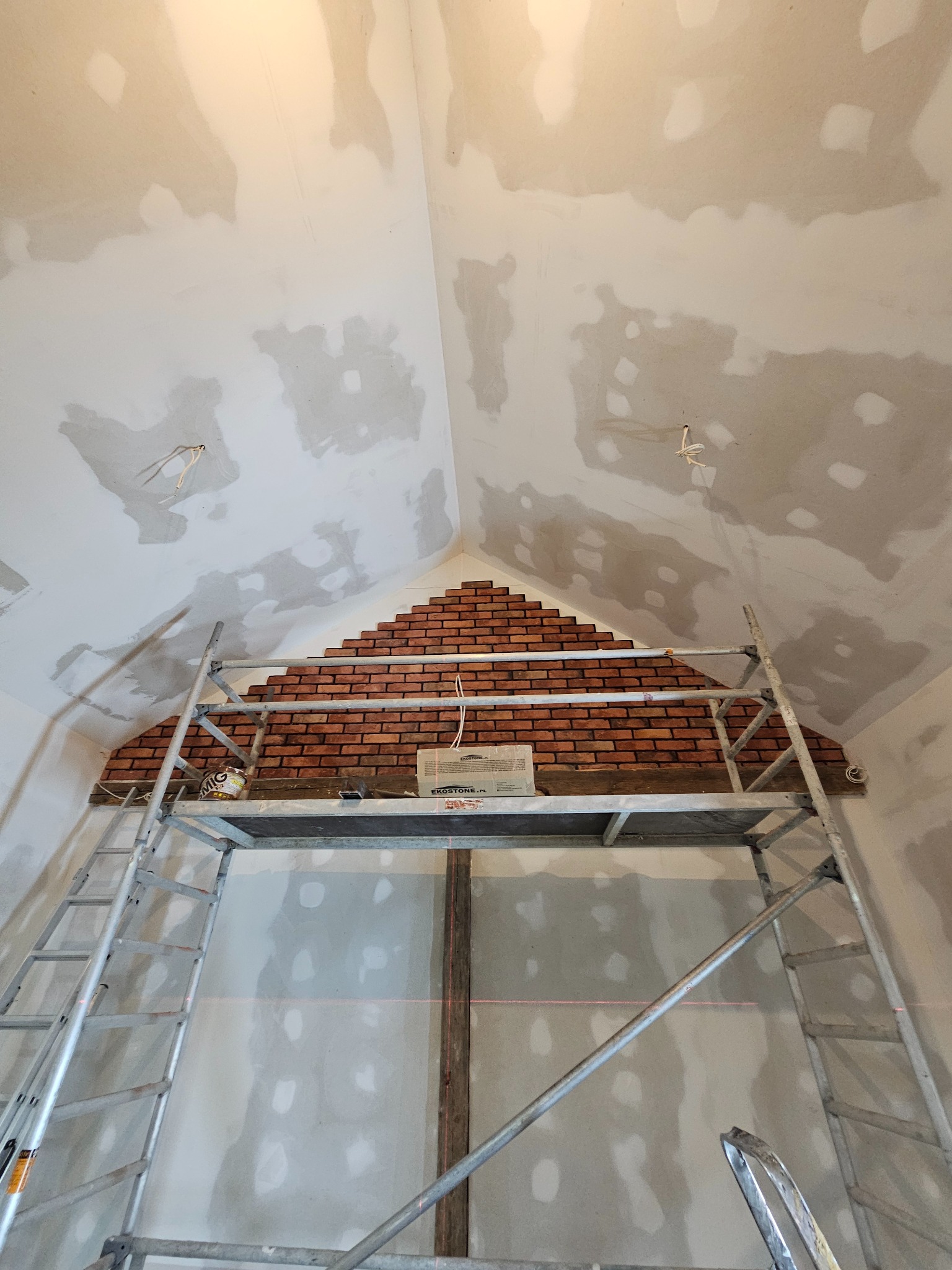 Montaż dekoracyjnej cegły na poddaszu, widoczny rusztowanie, surowy stan ścian i sufitu przed wykończeniem, czerwona cegła ułożona w trójkątny wzór, pudełko z materiałem budowlanym na rusztowaniu.