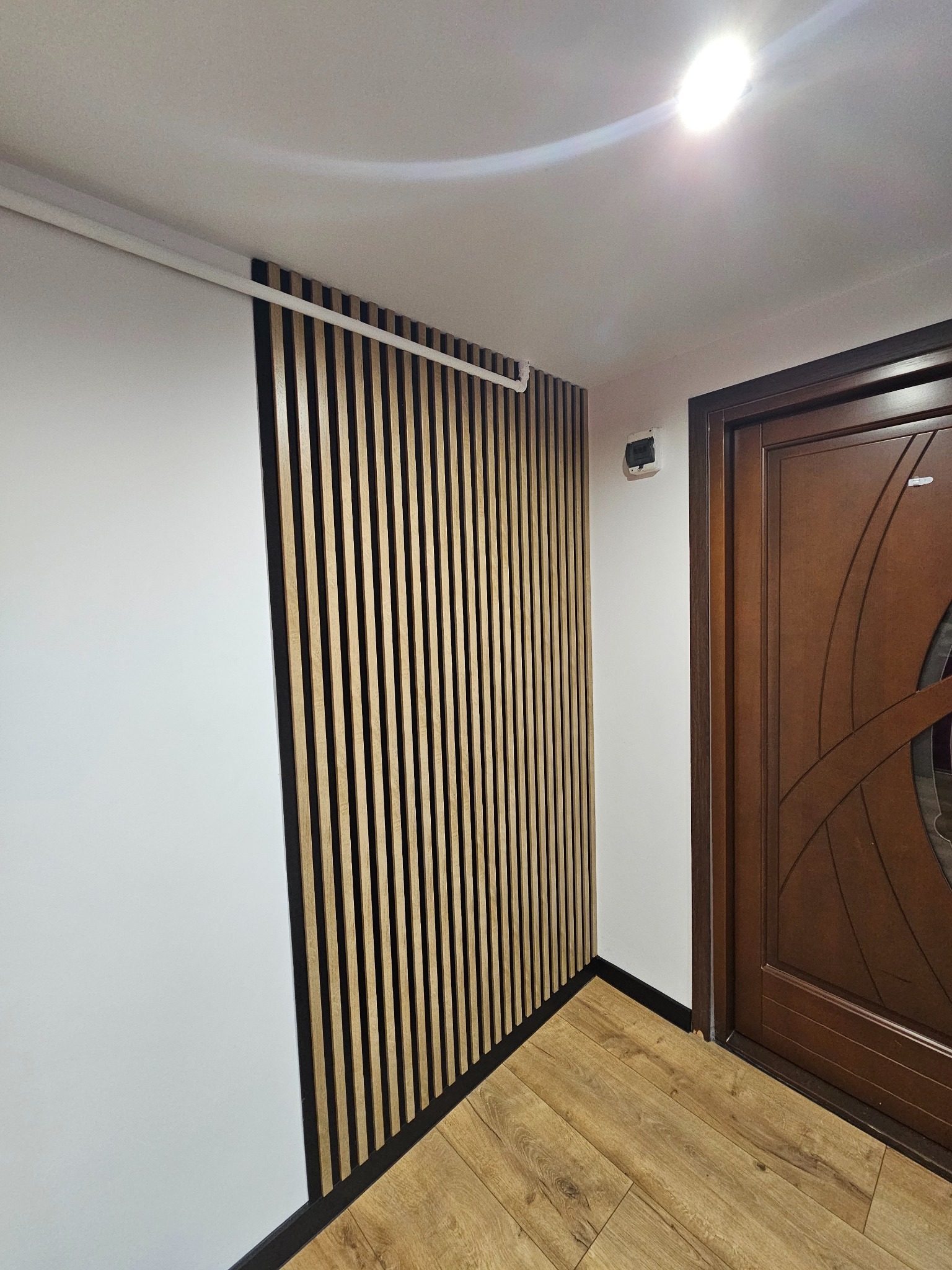 Panel z drewnianych listewek na ścianie, sąsiadujący z białą ścianą i brązowymi drzwiami, widoczne także panele podłogowe.