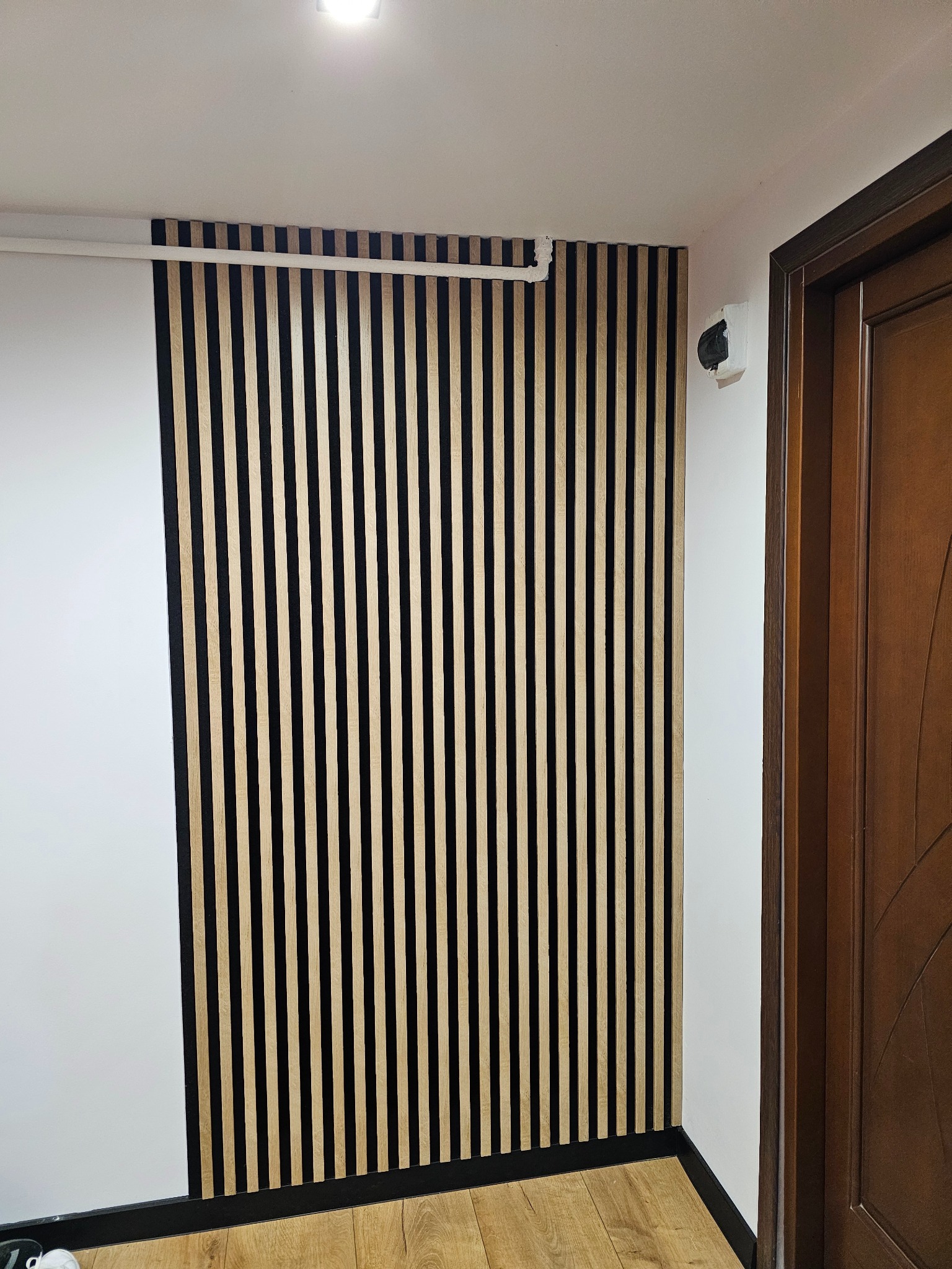 Nowoczesna ściana z drewnianych lameli w kolorze jasnego drewna, oddzielona od białej ściany czarną listwą, widoczna przy wejściu do pomieszczenia z drewnianą podłogą i framugą drzwi.