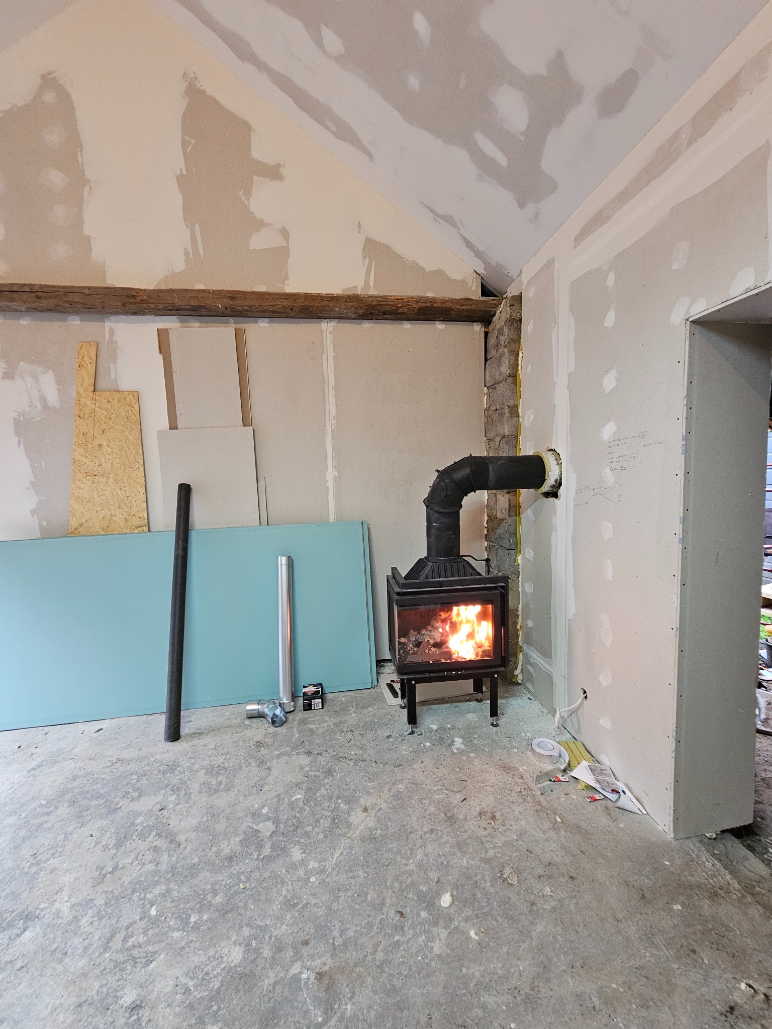 Wnętrze w trakcie remontu z widocznym kominkiem typu koza, płytami karton gips, surowym betonem na podłodze i drewnianą belką stropową.