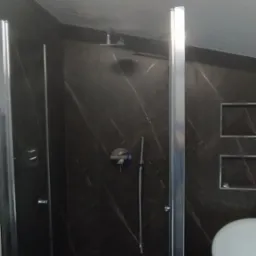 Szklana kabina prysznicowa z czarnymi marmurowymi płytkami, deszczownica i bateria podtynkowa, fragment umywalki widoczny po prawej stronie.